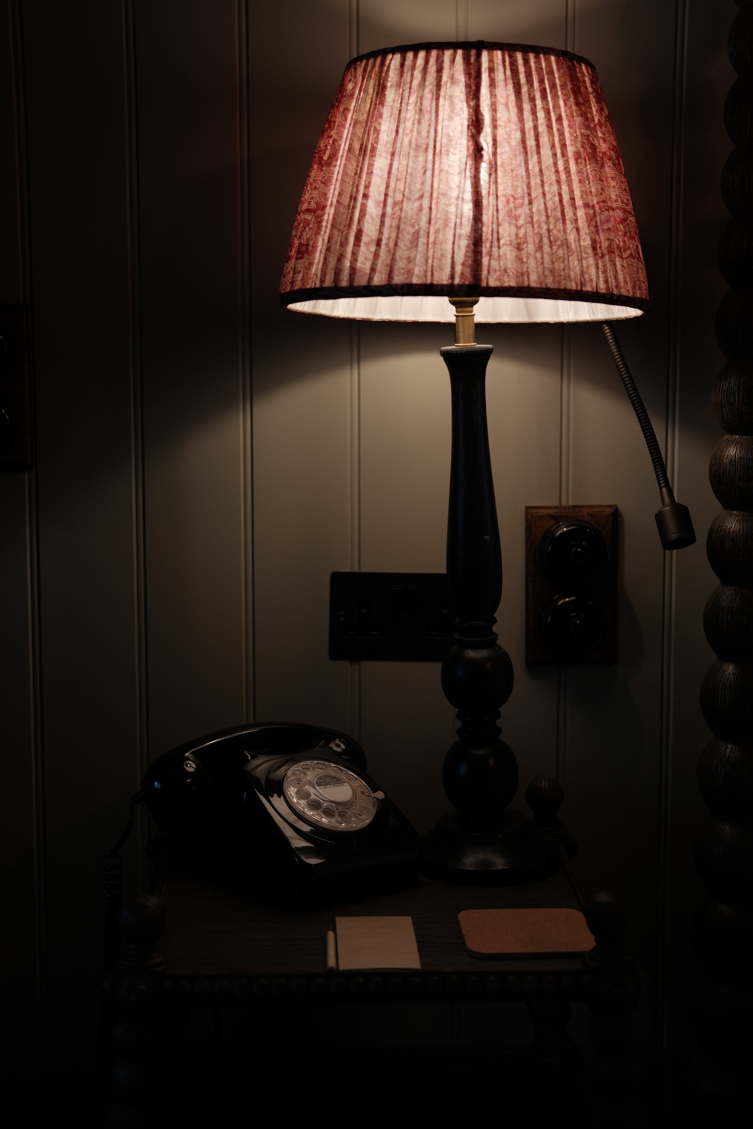 A vintage black rotary telephone on a dark wooden table, illuminated by a tall table lamp with a textured reddish-beige lampshade, against a paneled wall background.