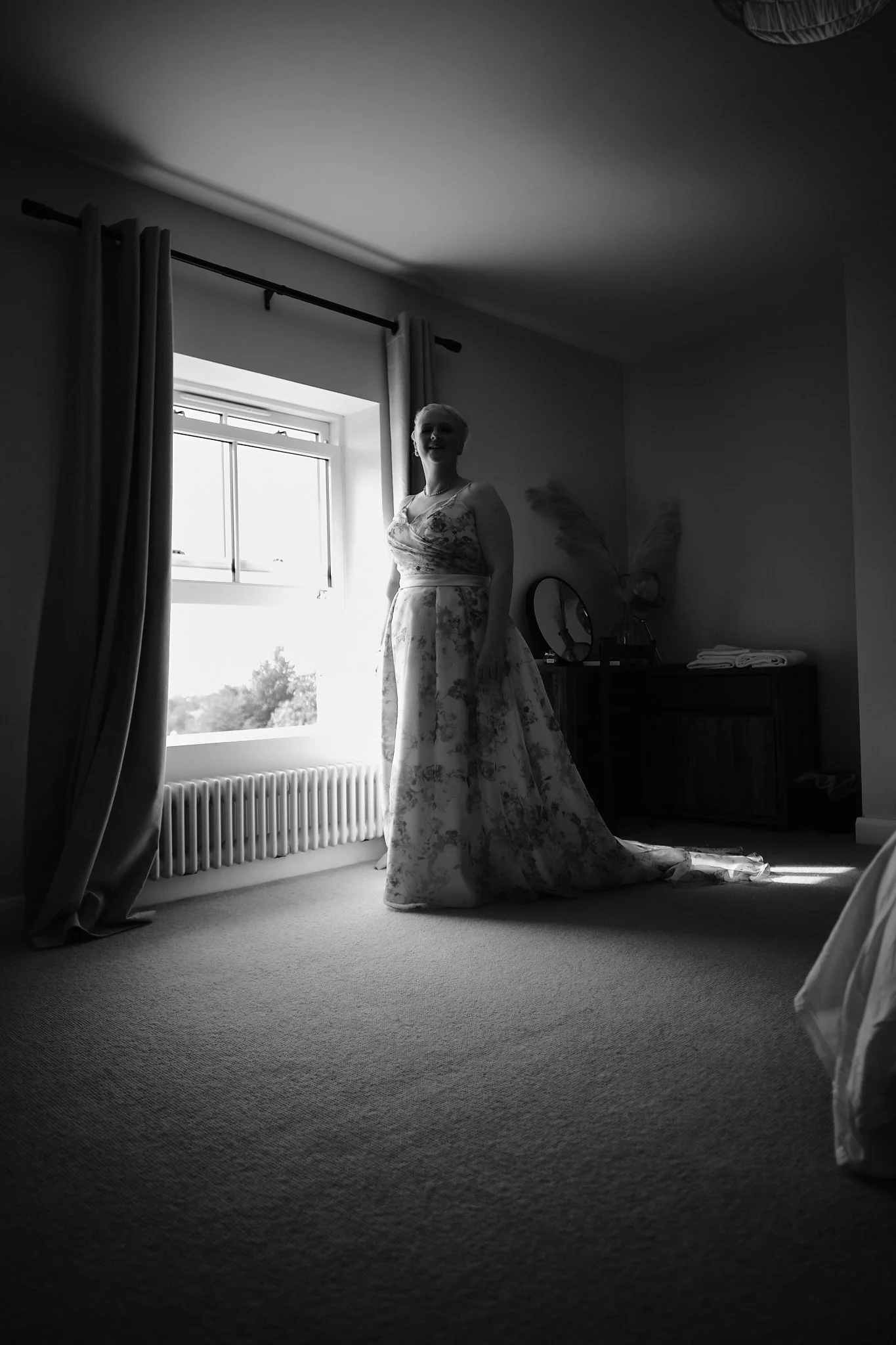 A woman in a floral wedding dress standing in a room with a large window, curtains, and a dresser, illuminated by natural light.