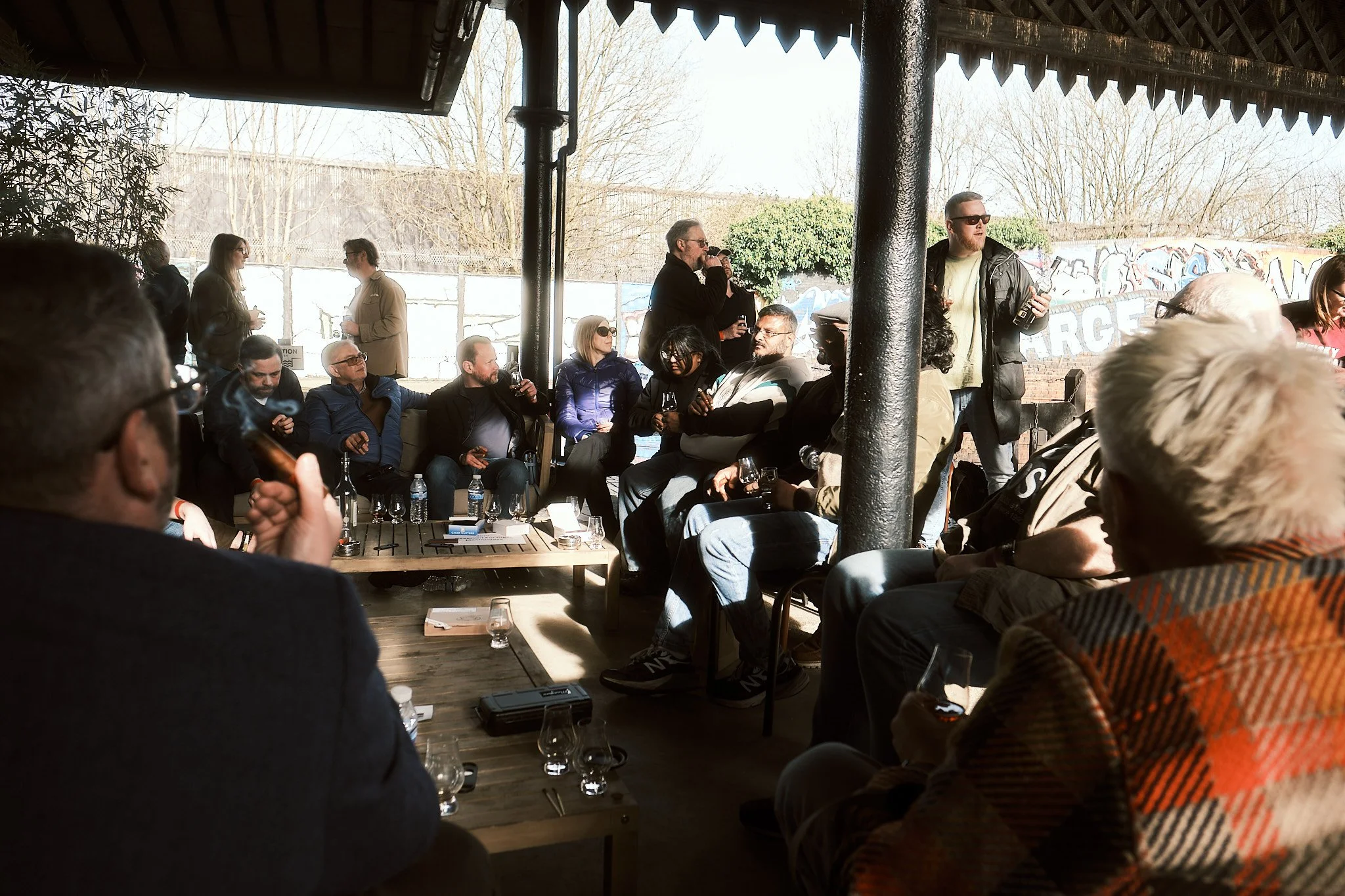 A group of people gathered in an outdoor patio area, some sitting and some standing, engaged in conversation and drinking beverages on a sunny day. The patio has a roof and there is graffiti on the wall in the background.