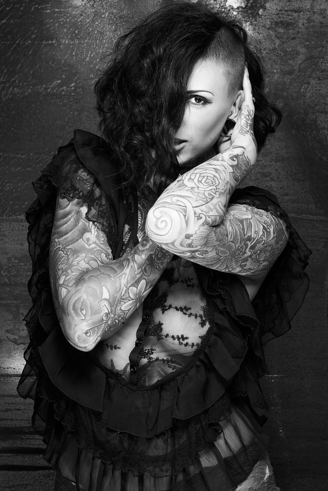 Black and white photo of a woman with tattoos, one side of her head shaved, posing with her face partially covered by her hand.