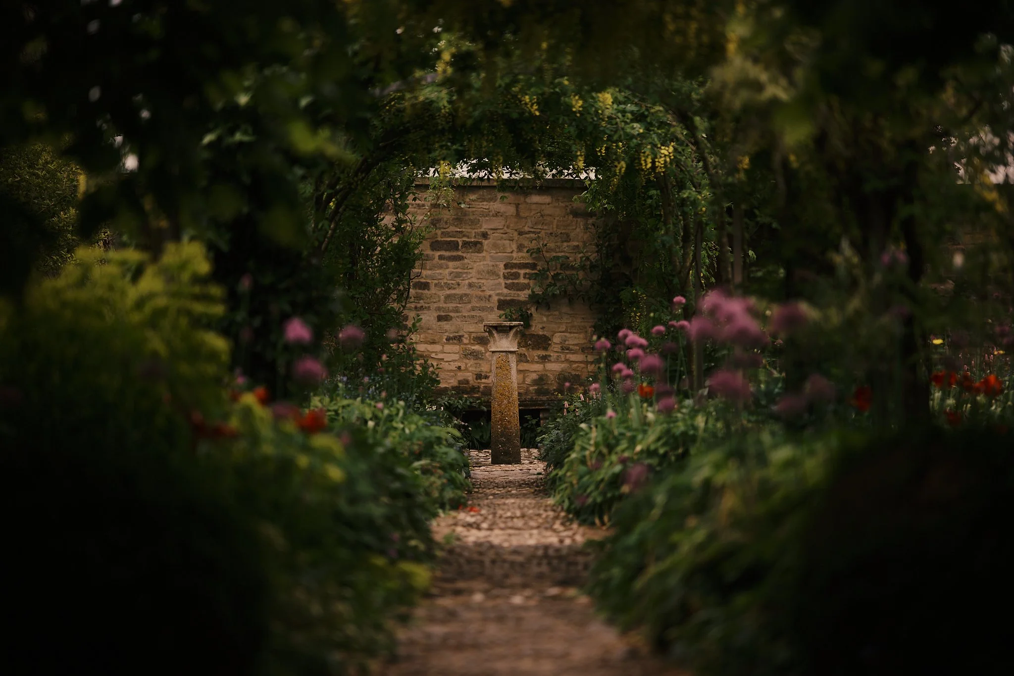 A garden pathway with a fountain in front of a stone wall, surrounded by bushes and colorful flowers, under an archway of greenery. Hotel interior photography at THE PIG-in the Cotswolds