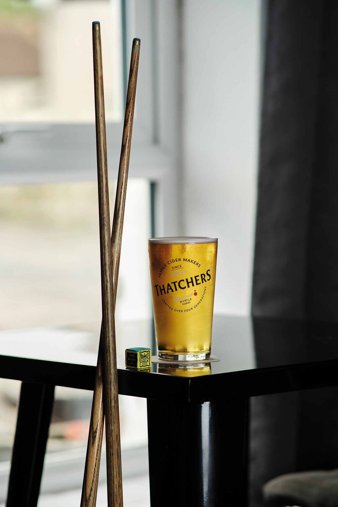 A pint glass of Thatcher's cider on a black table with a window in the background and two wooden chopsticks leaning against each other.