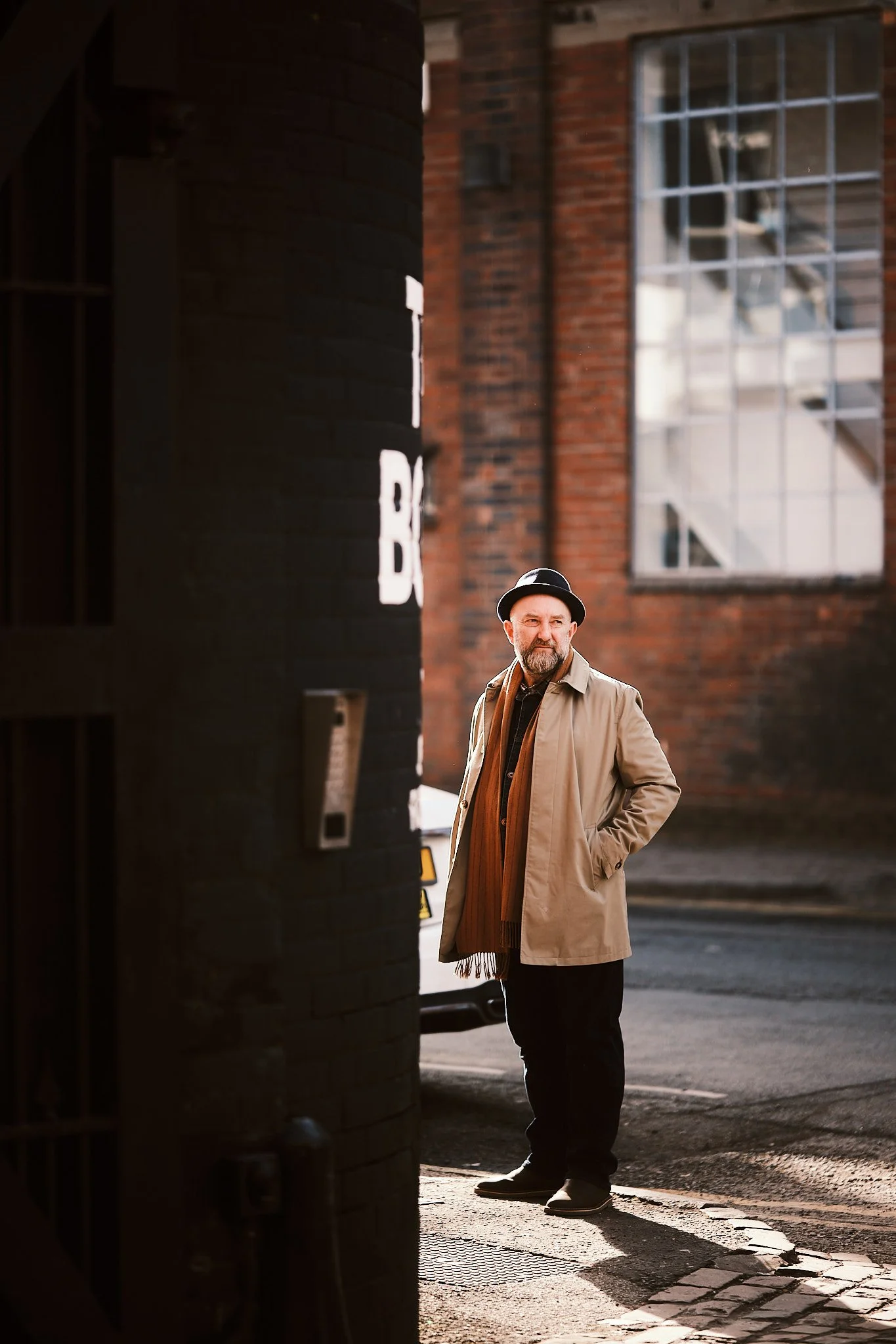 A man stands on a city sidewalk beside a brick building, wearing a beige coat, brown scarf, black hat, and black pants, with his hands in his pockets, during daytime.