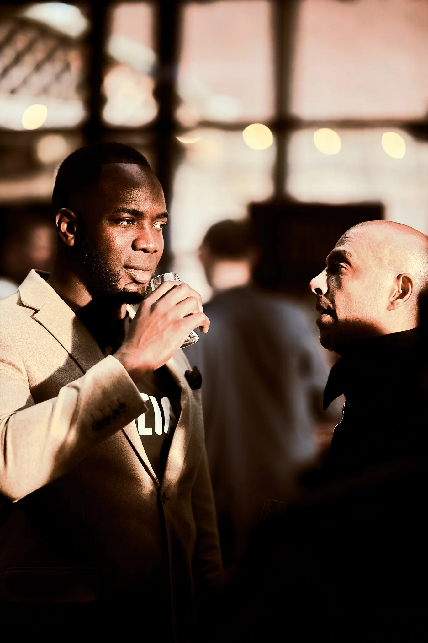 Two men conversing at a social event, one holding a glass, in a warmly lit indoor setting with blurred background.