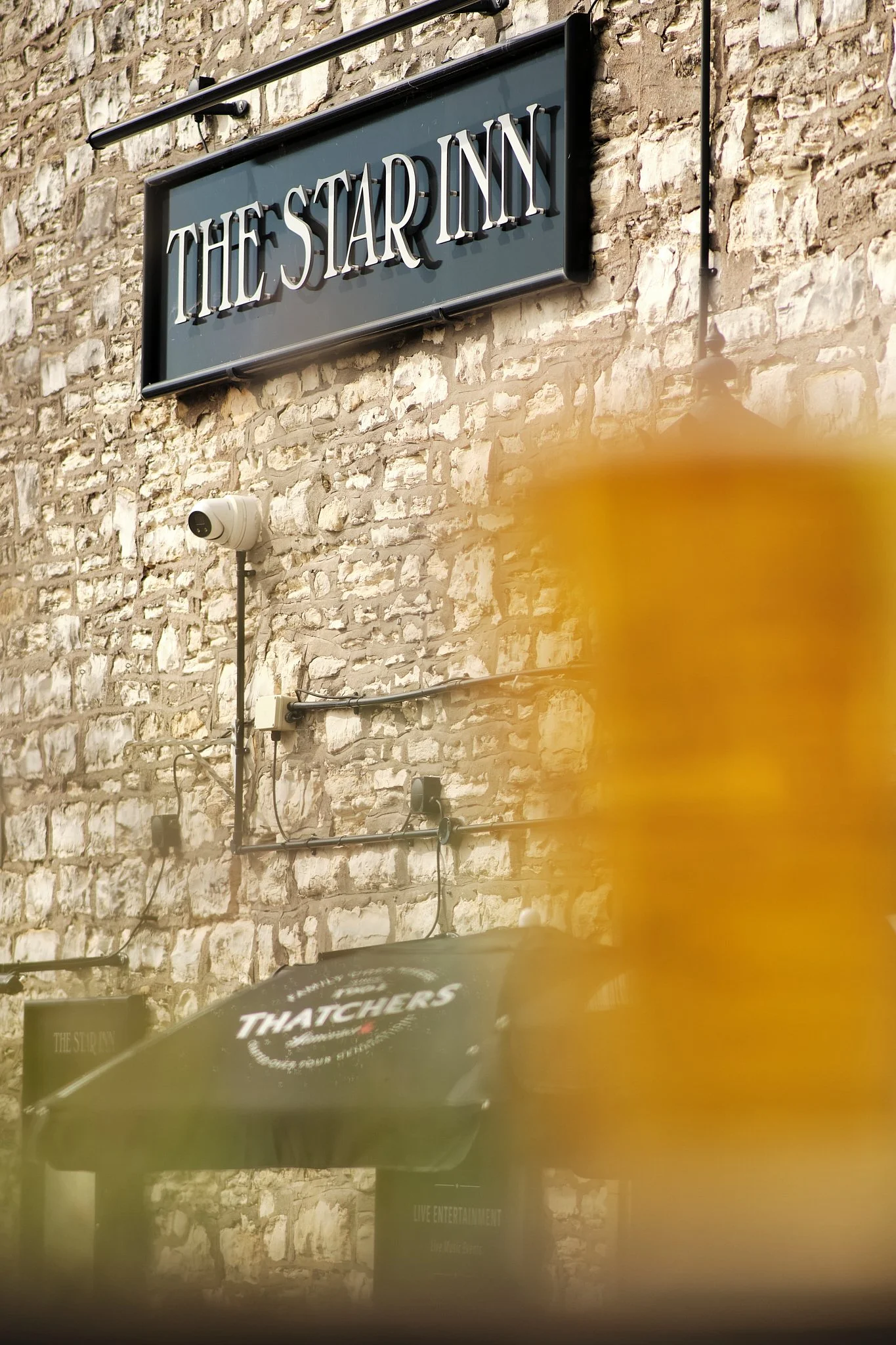 Sign for The Star Inn and outdoor seating with Thatchers logo umbrella, stone building exterior with security camera and lighting fixtures.