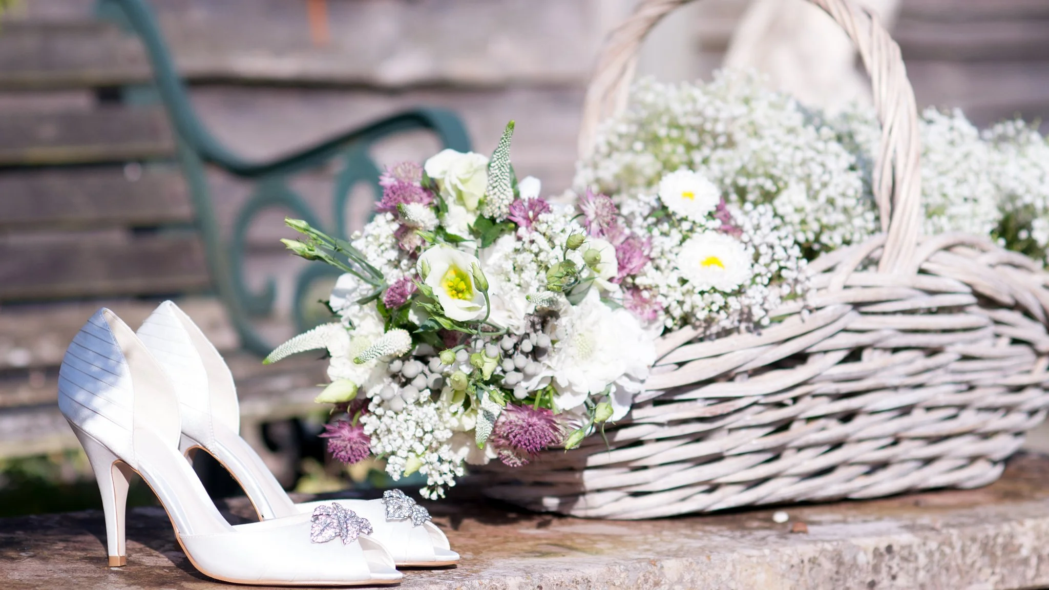 A pair of white high-heeled shoes with satin finish and bows on the toe, positioned next to a wicker basket filled with white and purple flowers, on a wooden surface.