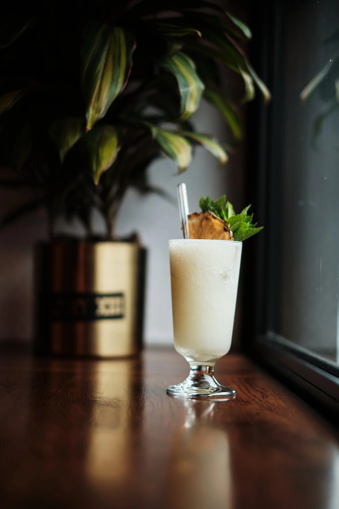 A tall, frothy, creamy cocktail in a glass garnished with a pineapple slice and mint leaves, placed on a wooden table near a window with a green plant in the background.