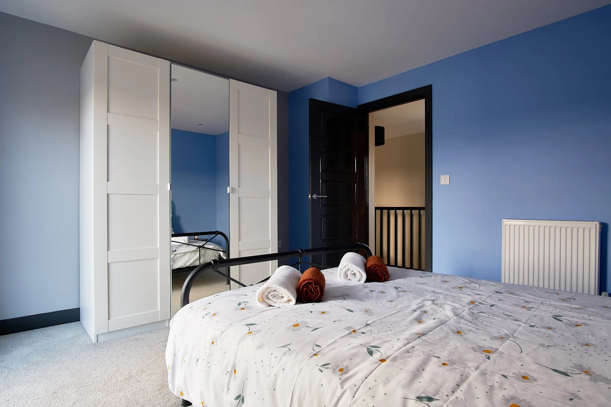 A bedroom with a blue wall, white wardrobe with mirrored doors, black bed frame, and a bed with white floral bedding and rolled towels on top.
