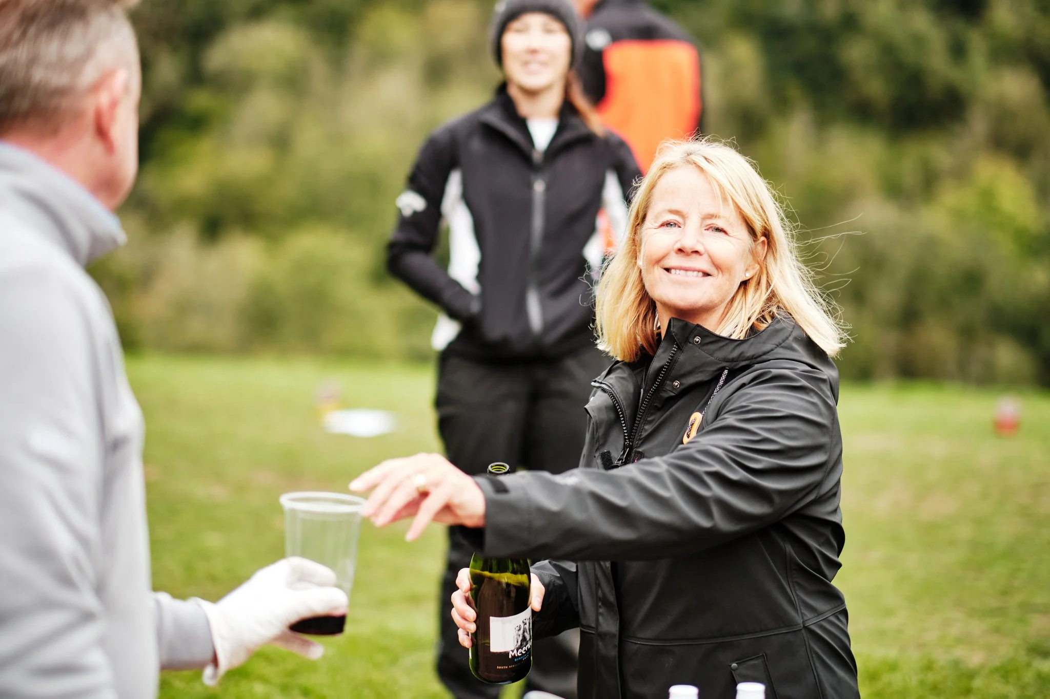 A woman in a black jacket is smiling and handing a glass of what appears to be red wine to another person during an outdoor gathering in a park-like setting, with another woman standing in the background.