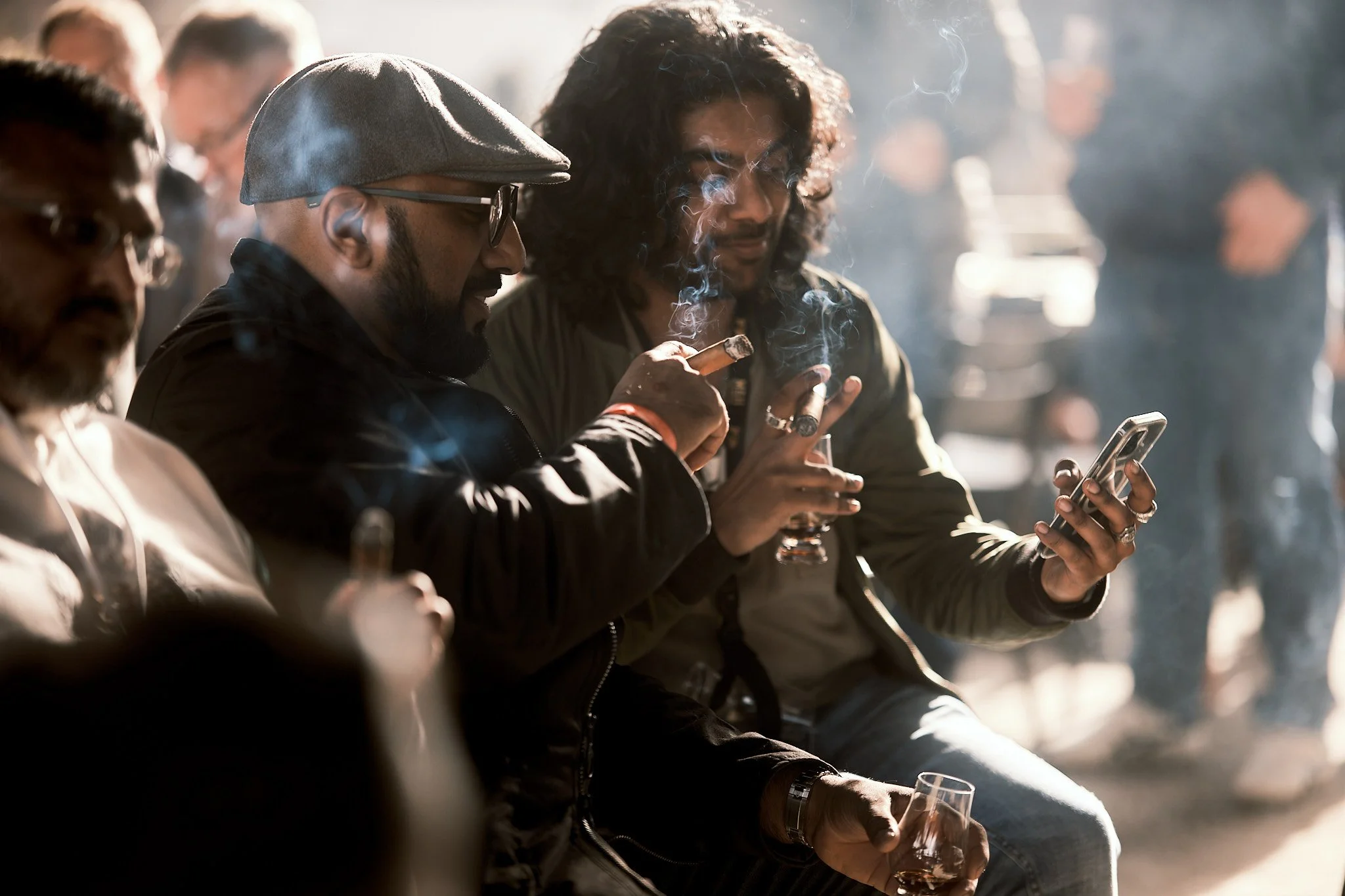 Three men sitting together, two of them smoking cigars and one looking at a smartphone, with others in the background.