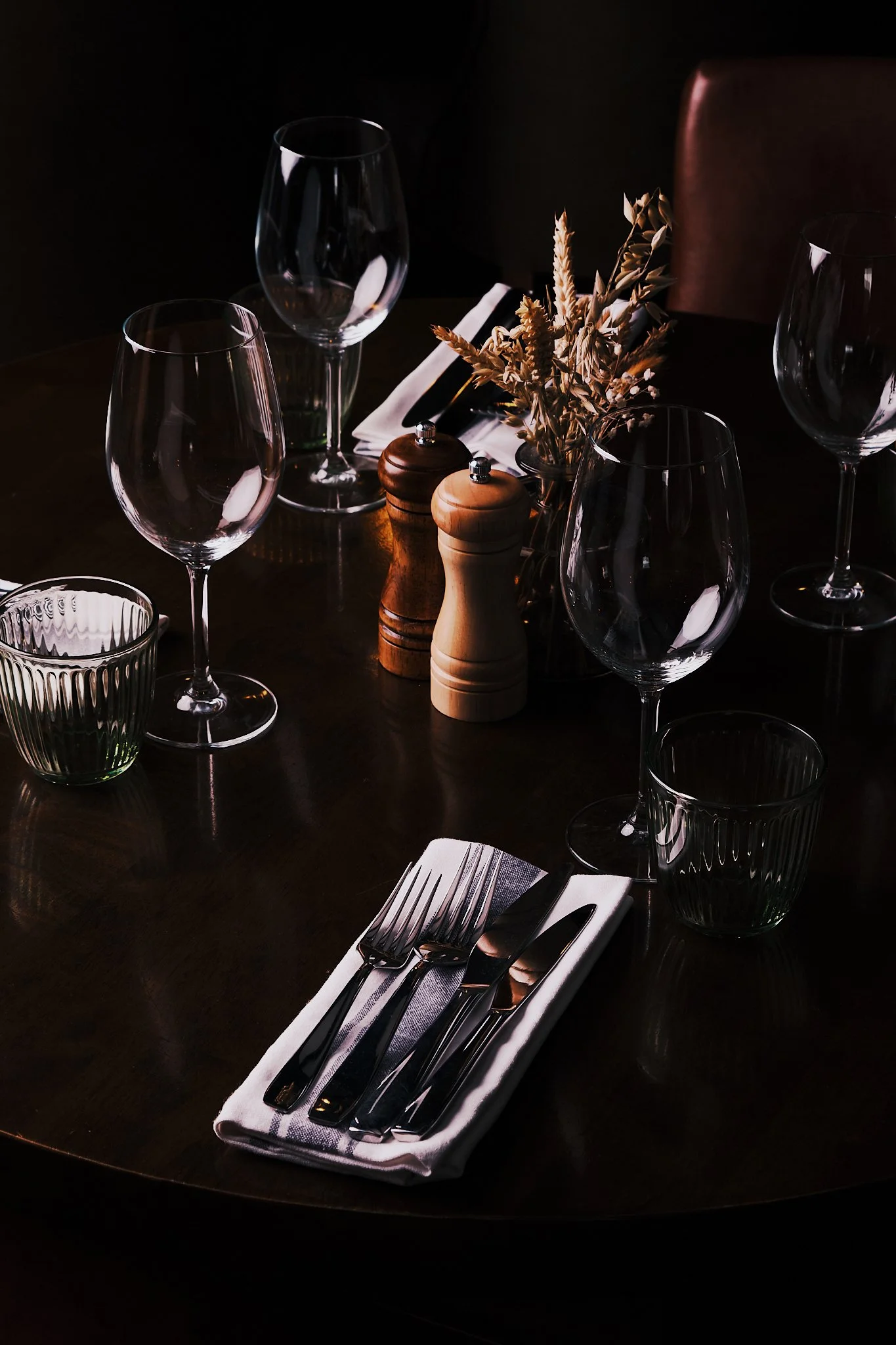 A dining table set with wine glasses, water glasses, cutlery, salt and pepper shakers, a folded napkin, and a vase with dried flowers in a dimly lit room.