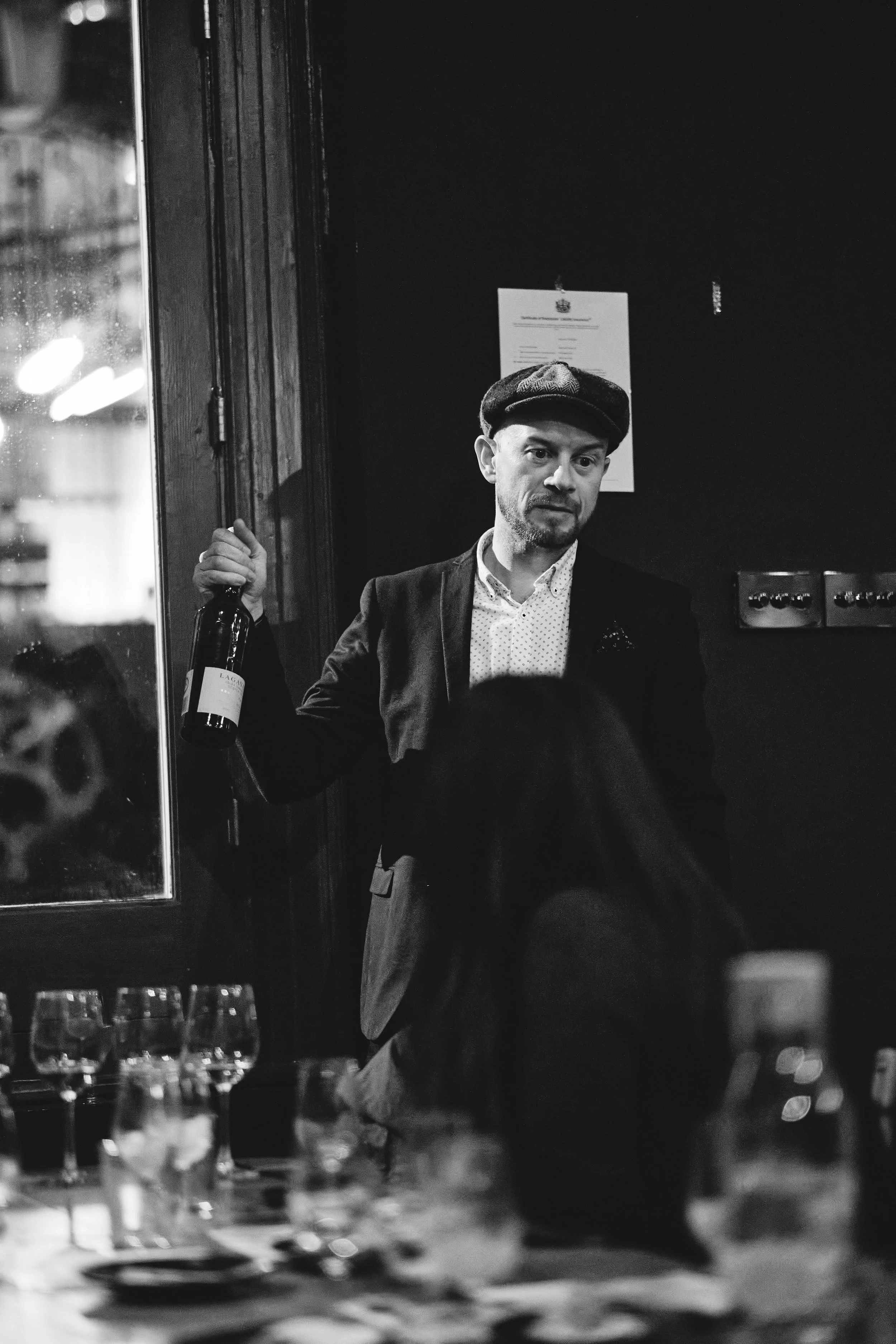 A man holding a wine bottle at a restaurant or bar, dressed in a blazer and wearing a flat cap, in a black and white photo.