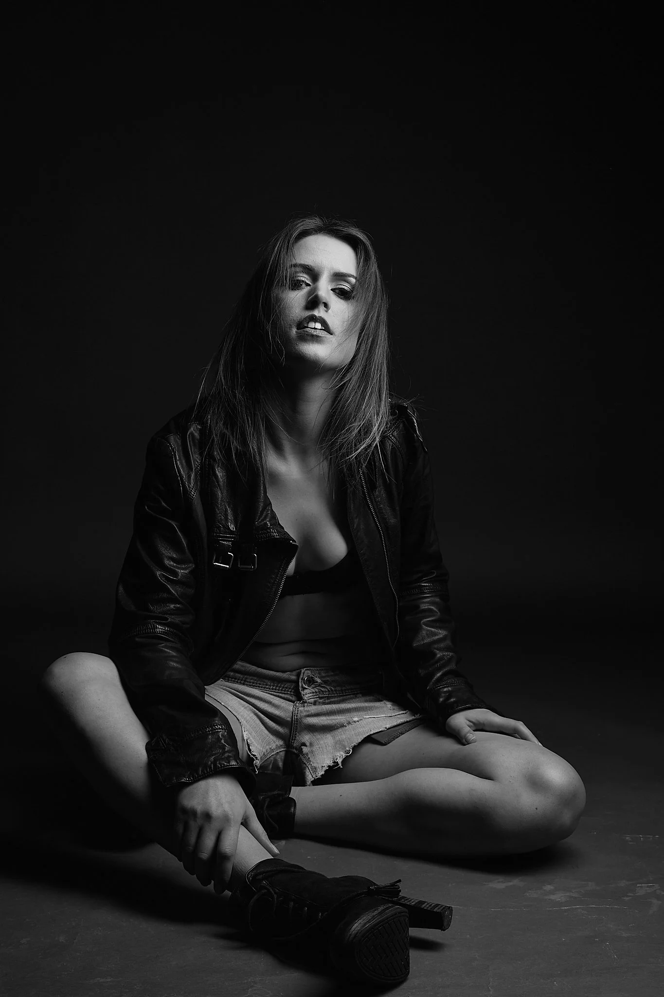 Black and white photo of a woman sitting on the floor with crossed legs, wearing a leather jacket, revealing her midriff, with messy hair, against a dark background.