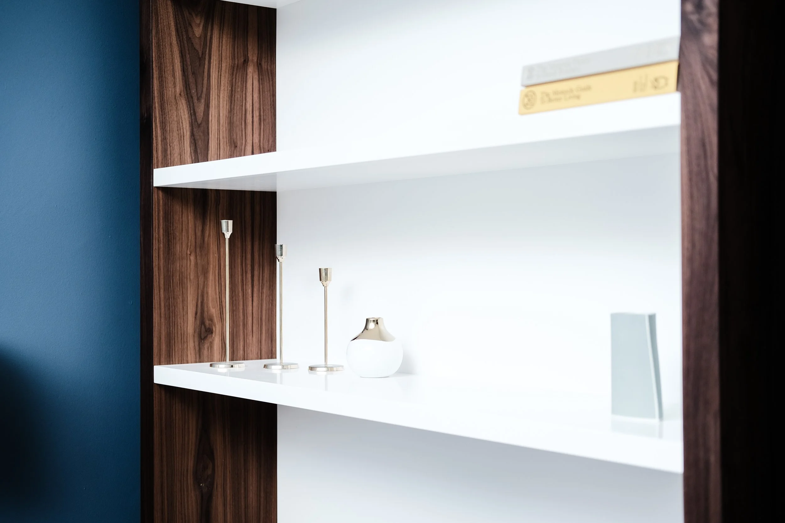 Empty white and dark wood bookshelf with three gold candlestick holders, a small white and gold decorative vase, and a few boxes on the upper shelves.