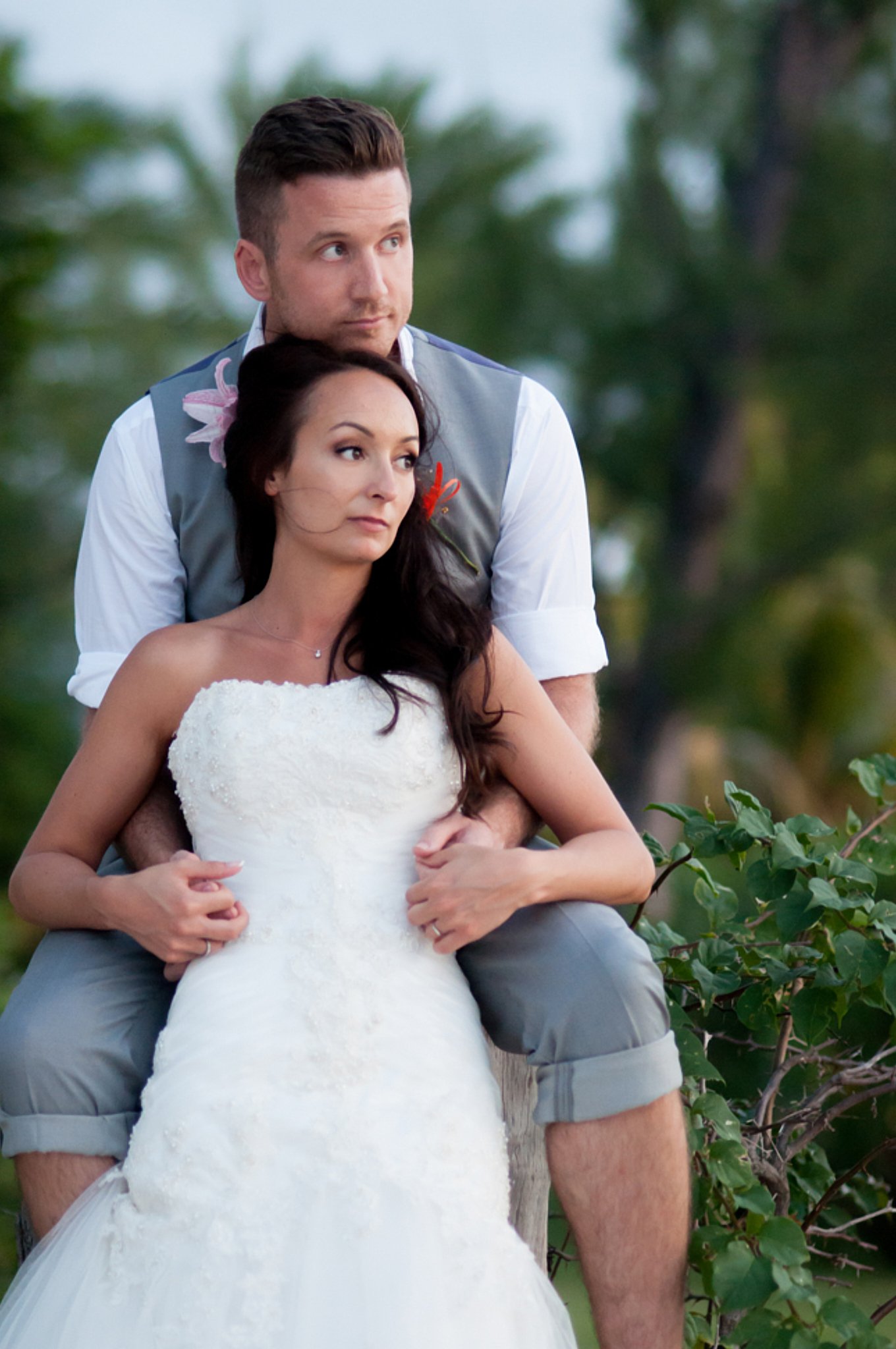 A newly married couple, a man in a gray vest and a woman in a white wedding dress, sitting outdoors with greenery in the background.
