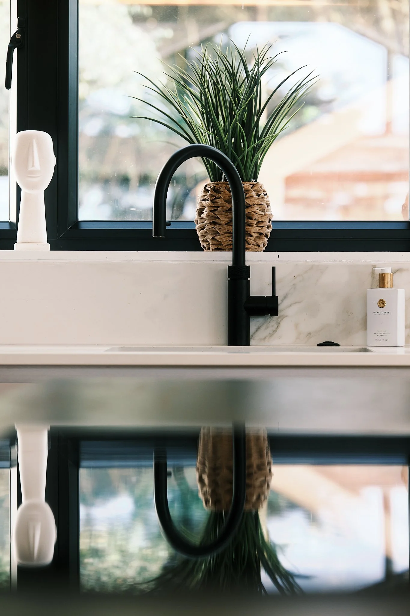 A kitchen sink area with a black faucet, a potted plant with long green leaves, a white decorative bust, and a bottle of soap, with a window in the background.