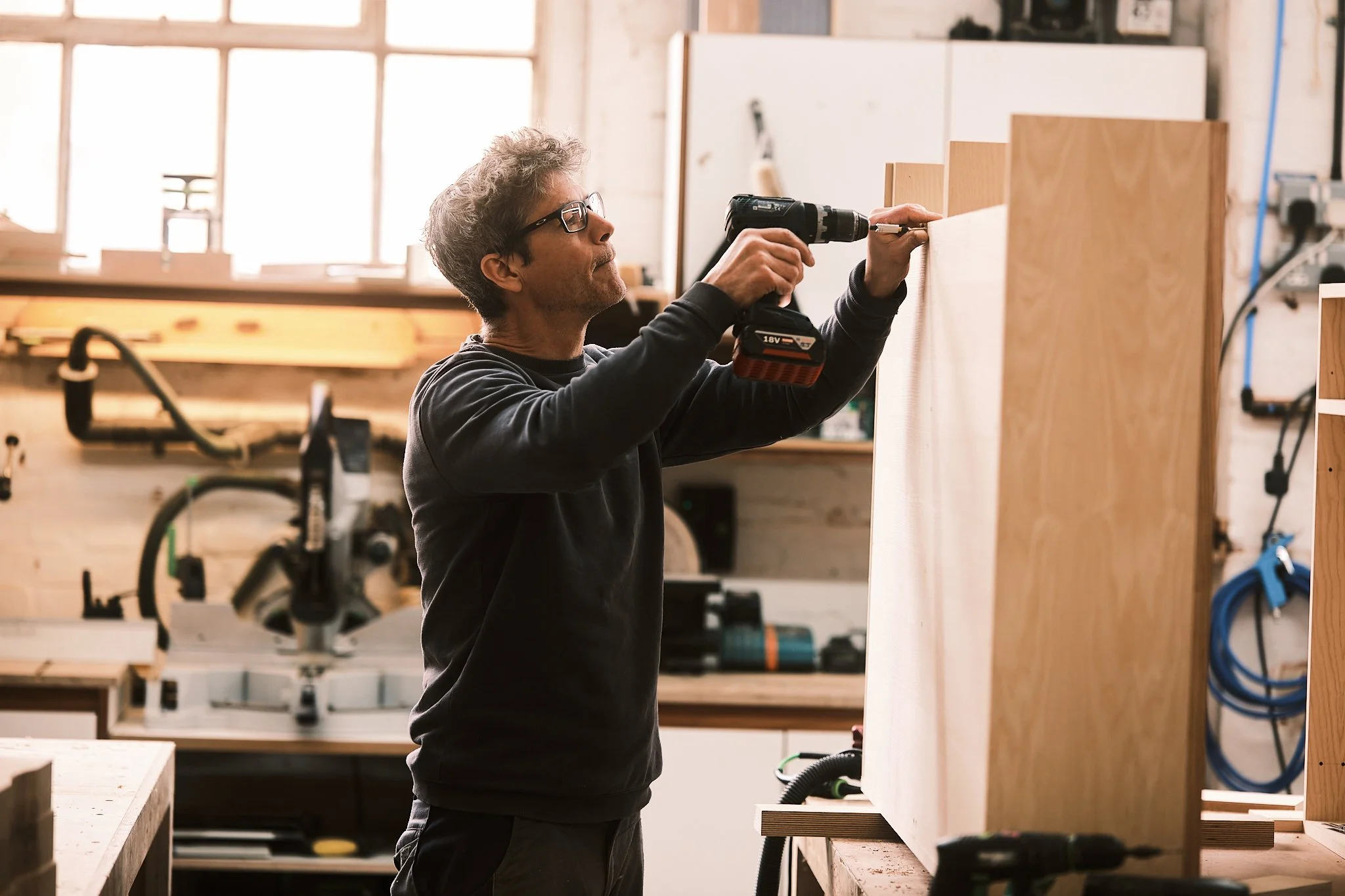 A man with gray hair and glasses using a cordless drill to work on a wooden panel in a woodworking shop.