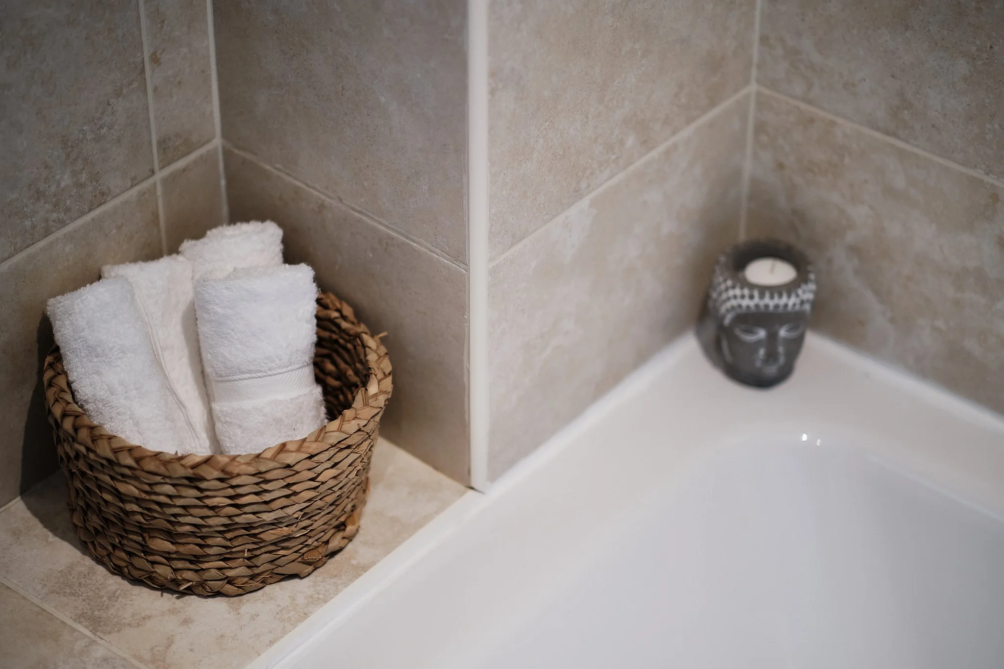 White towels folded in a wicker basket in a tiled bathroom corner, with a decorative candle holder featuring a face design on the bathtub ledge.