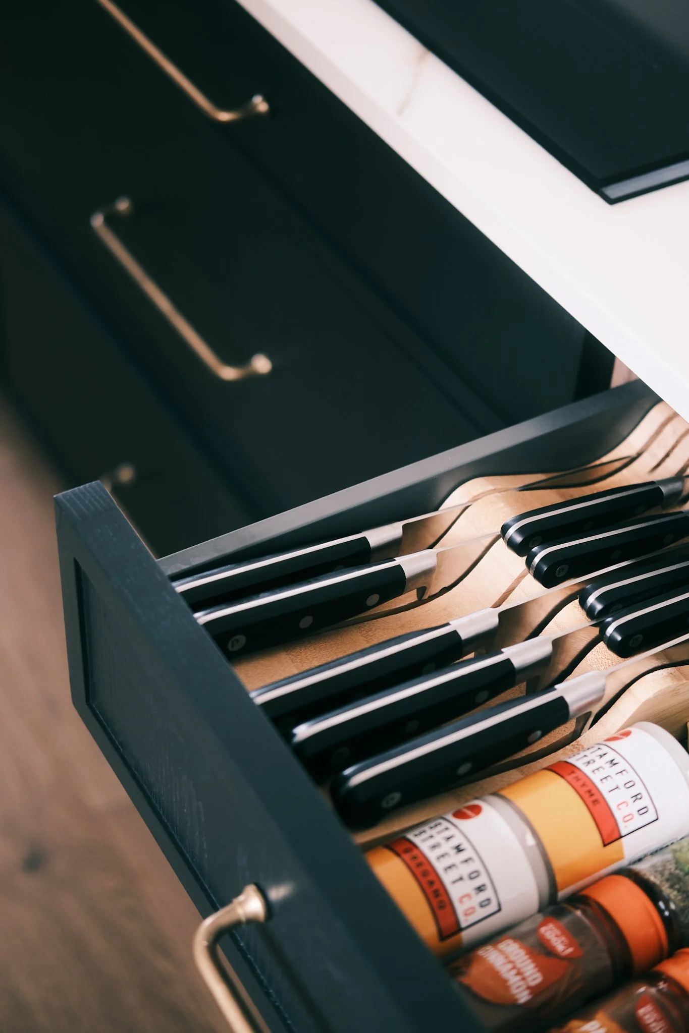 Drawer with black-handled knives, spices, and a black grill pan on a white countertop.