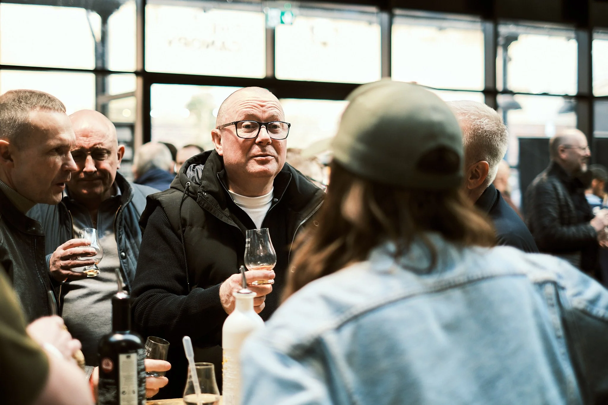 People socializing at a bar, holding drinks, in an indoor setting with large windows.