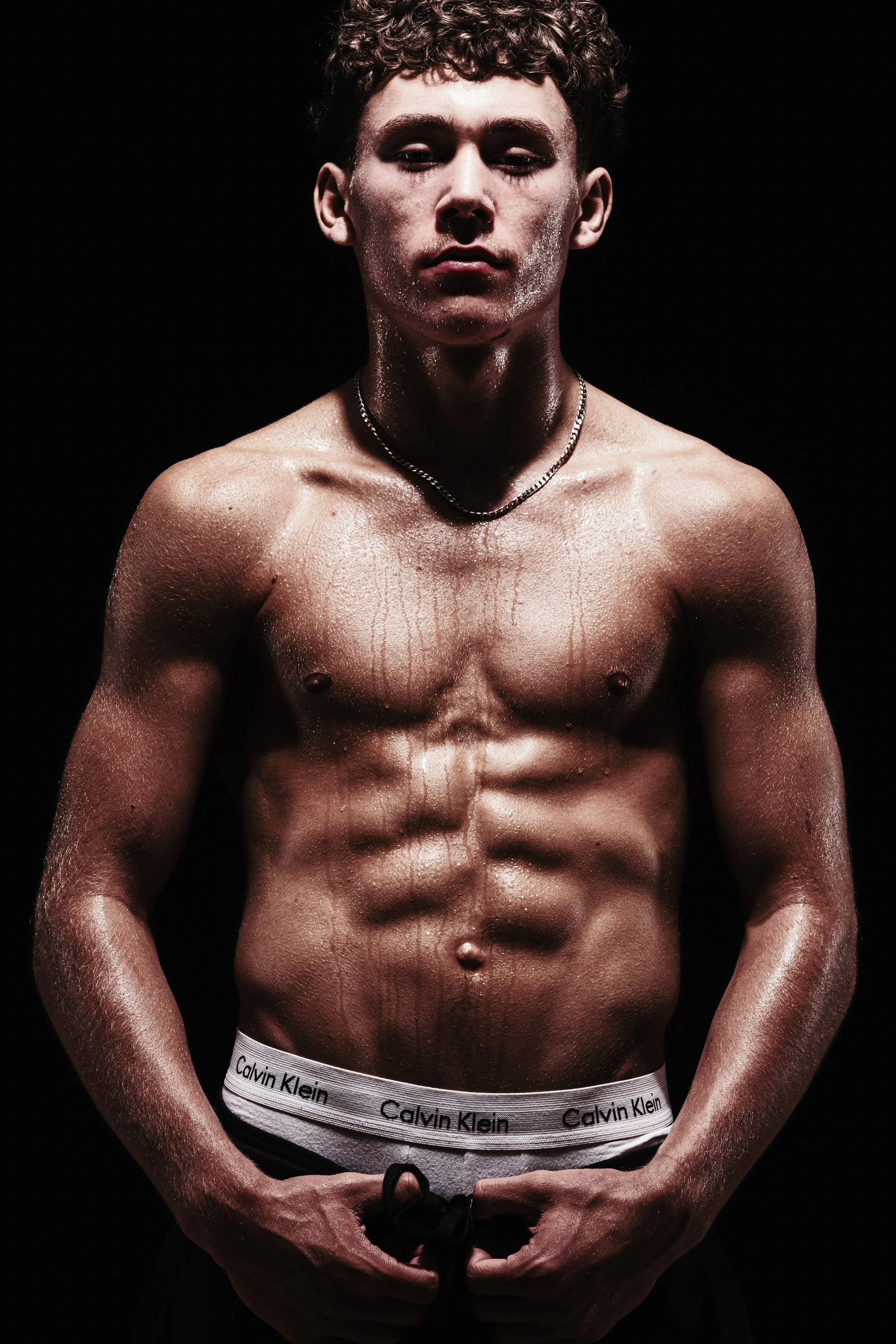 A shirtless young man with curly hair and a toned physique, standing against a black background. He is glistening with sweat and wears a chain necklace and Calvin Klein underwear.