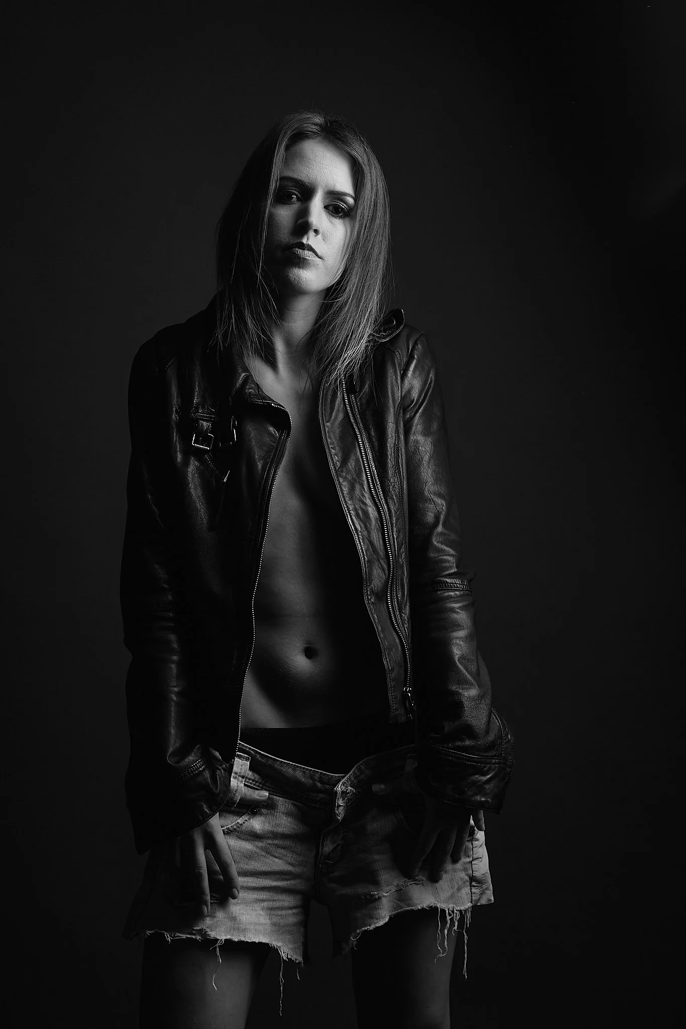 A black and white photo of a young woman with mid-length hair, wearing a leather jacket and ripped denim shorts, standing against a dark background.
