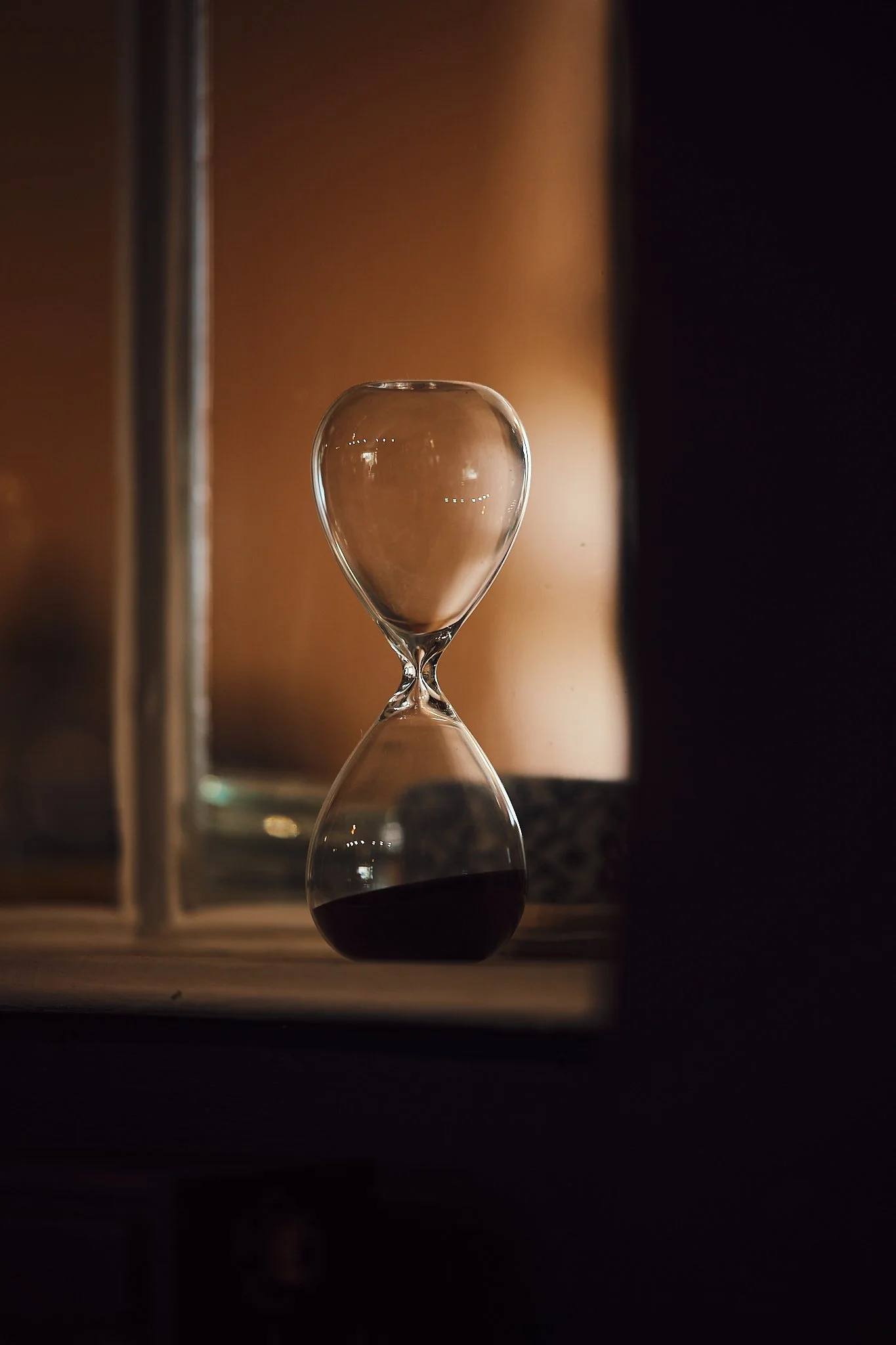 An hourglass with black sand half full, positioned in front of a mirror in a dimly lit room.