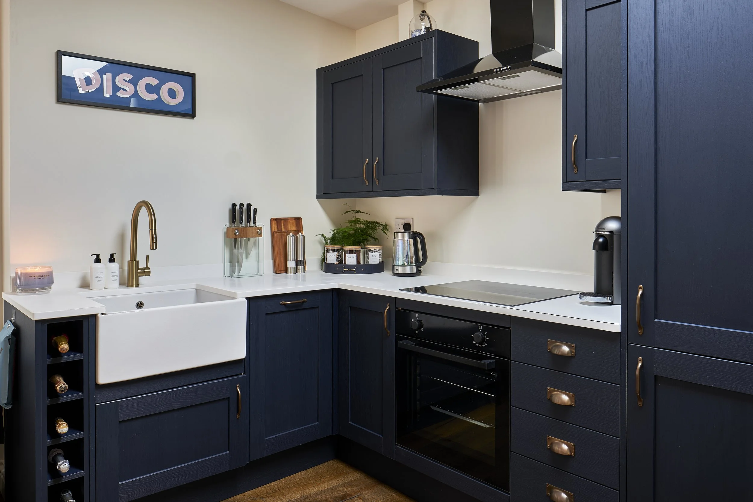 Kitchen with dark blue cabinets, white countertop, farmhouse sink, and black oven. Decor includes a 'DISCO' sign on the wall, knife holder, plants, and small appliances.