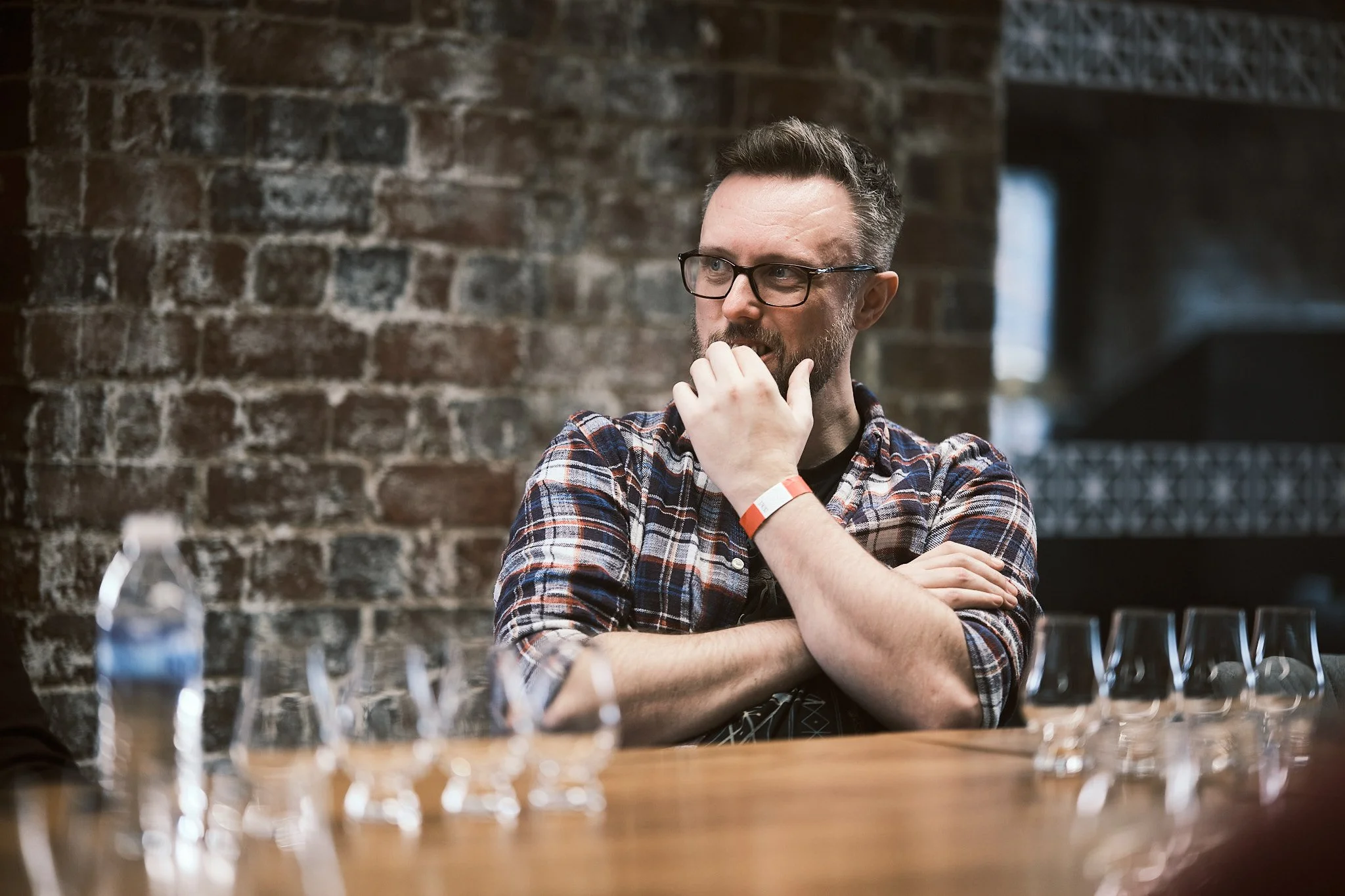 A man with glasses, wearing a plaid shirt, sitting at a table with several glasses and a water bottle, in front of a brick wall.