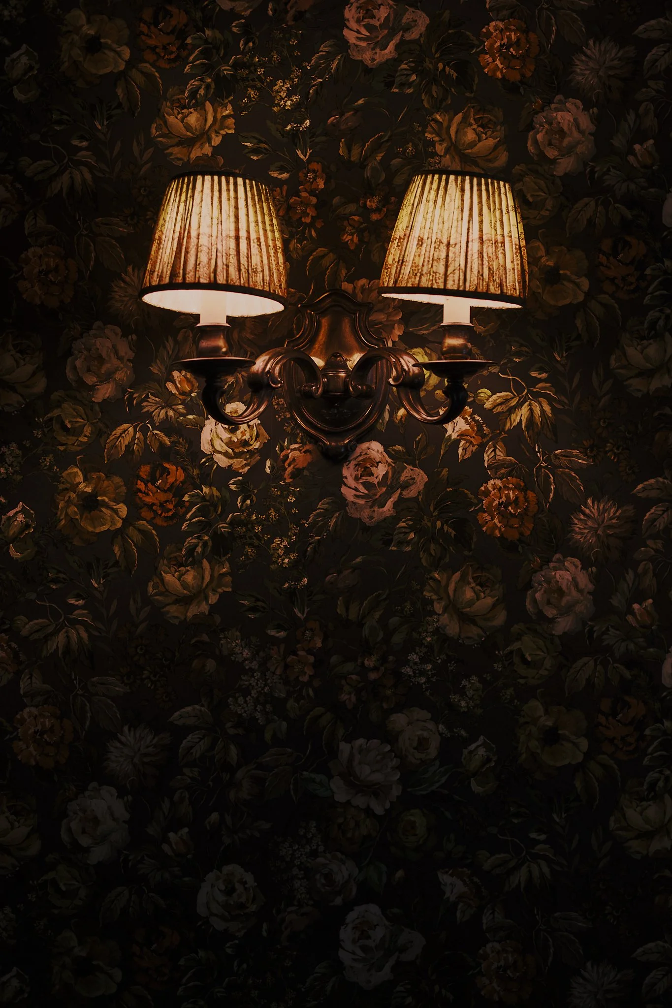A dark floral wallpaper with a vintage-style wall sconce light fixture featuring two lampshades emitting a warm glow. Hotel interior photography at THE PIG-in the Cotswolds