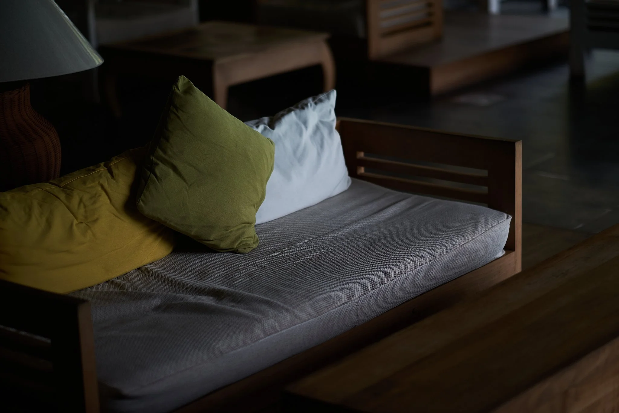 A wooden daybed with a light gray cushion, three pillows (green, yellow, and white), and a wooden table in a dimly lit room.