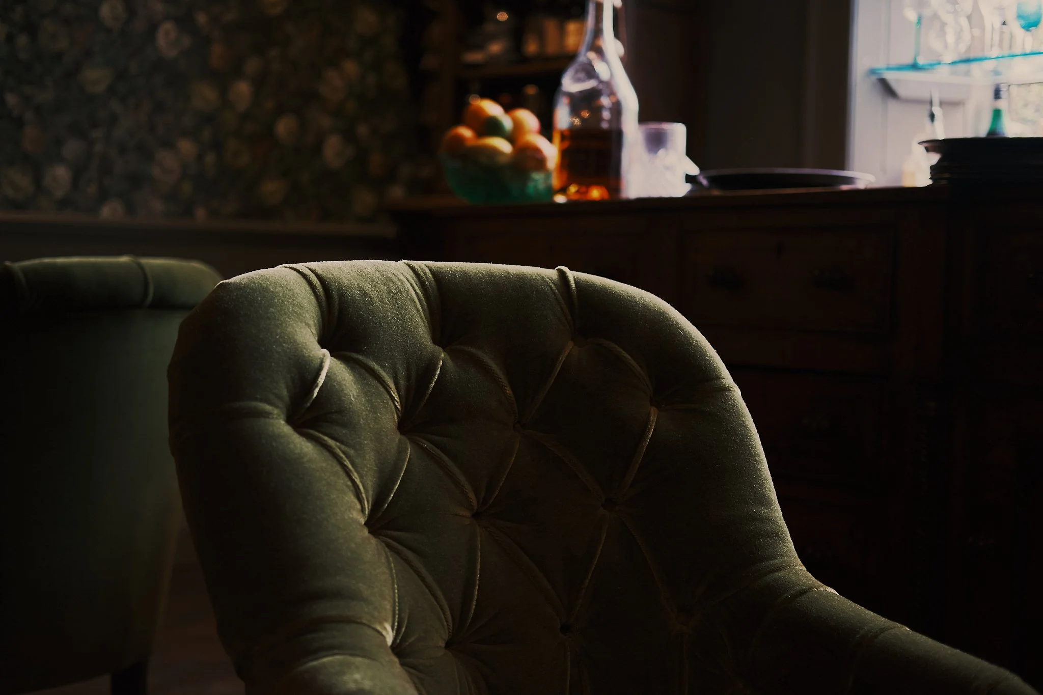 Close-up of a vintage green velvet armchair in a dimly lit room, with a wooden sideboard and a window with visible glass and a bottle and fruit on the sideboard in the background. Hotel interior photography at THE PIG-in the Cotswolds