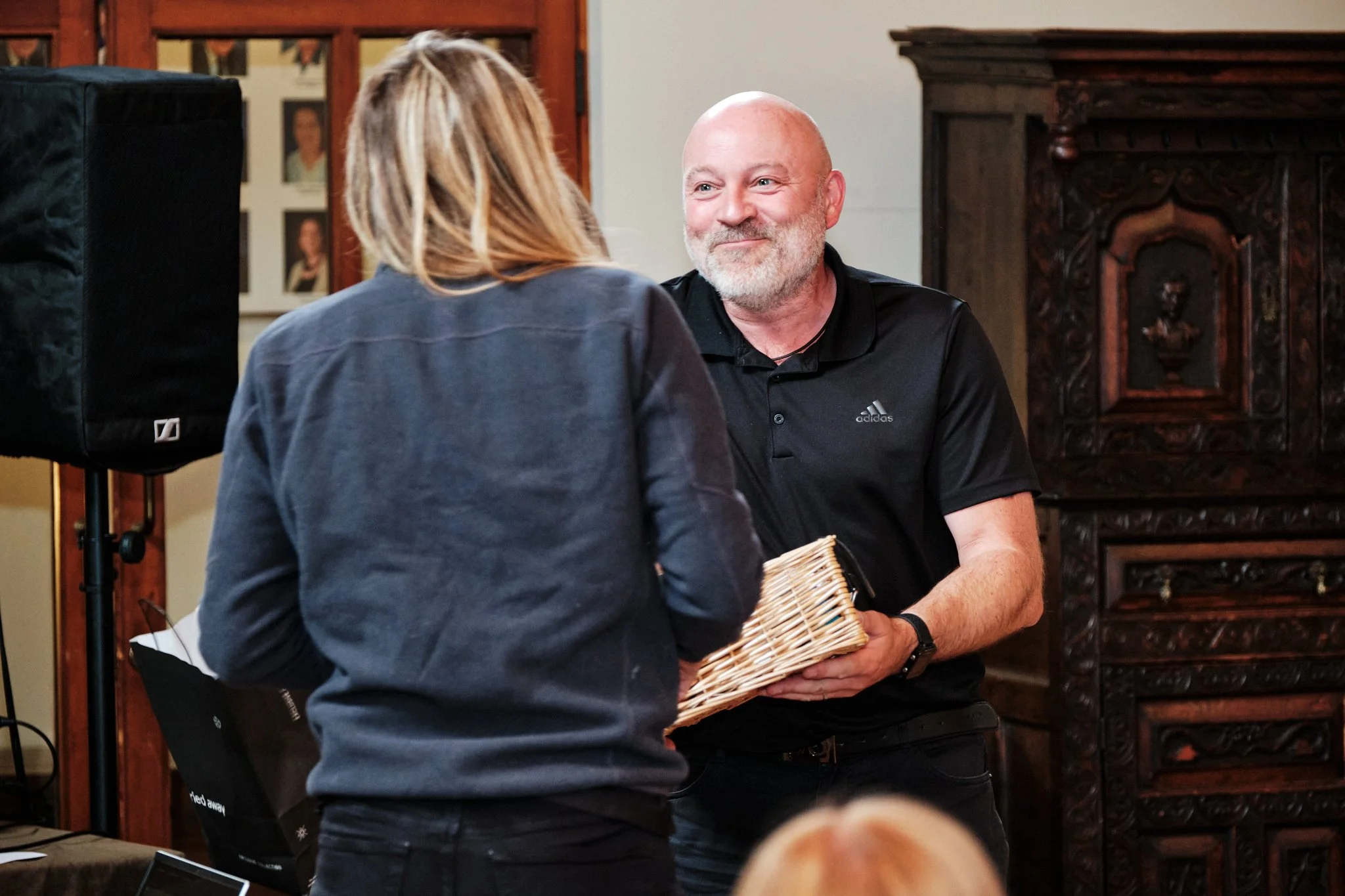 A man with a beard, wearing a black Adidas shirt, is smiling while holding a wicker basket, and he is interacting with a woman with shoulder-length blonde hair, wearing a dark jacket, in a room with wooden furnishings.