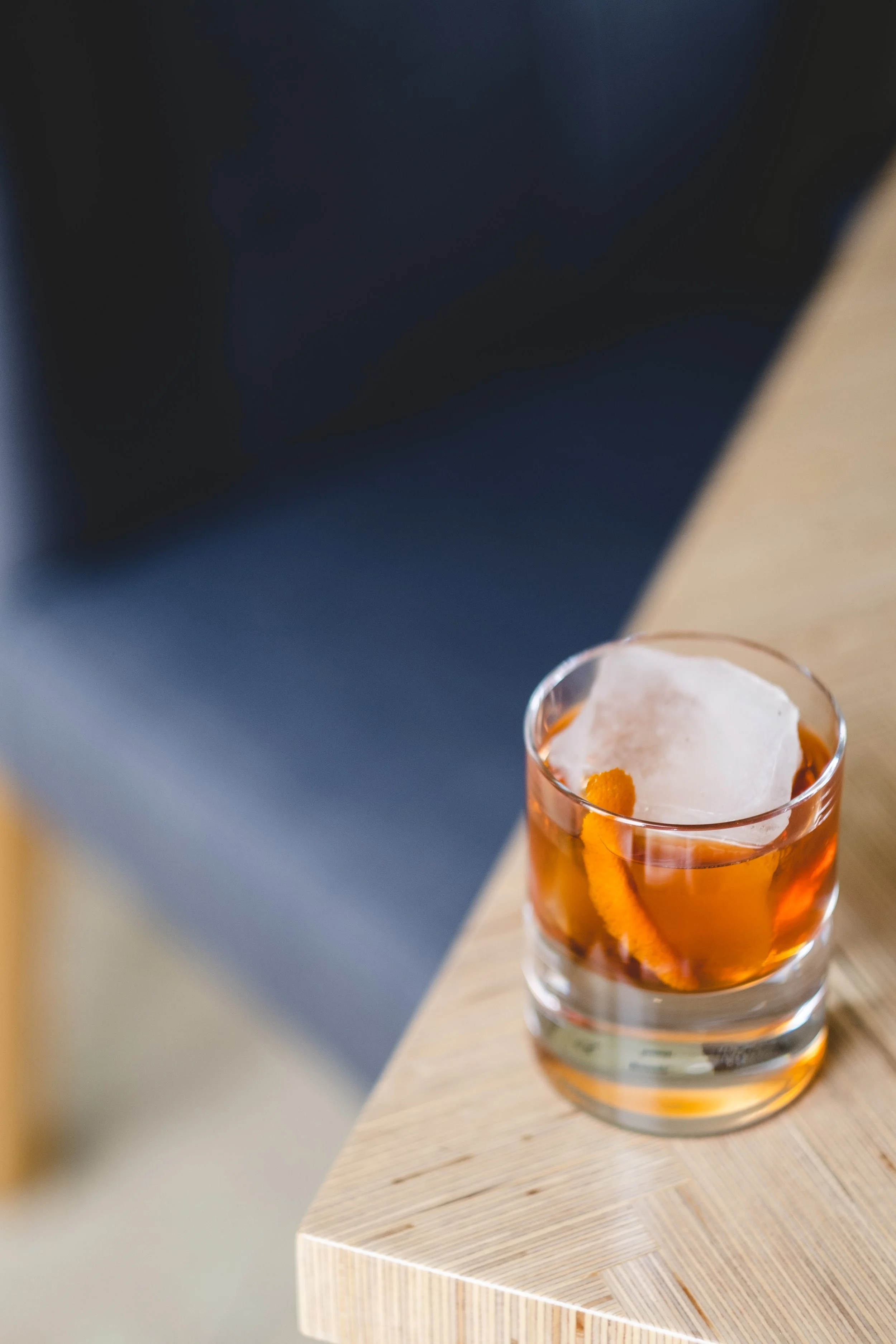 A glass of whiskey with orange peel and a large ice cube on a wooden table.