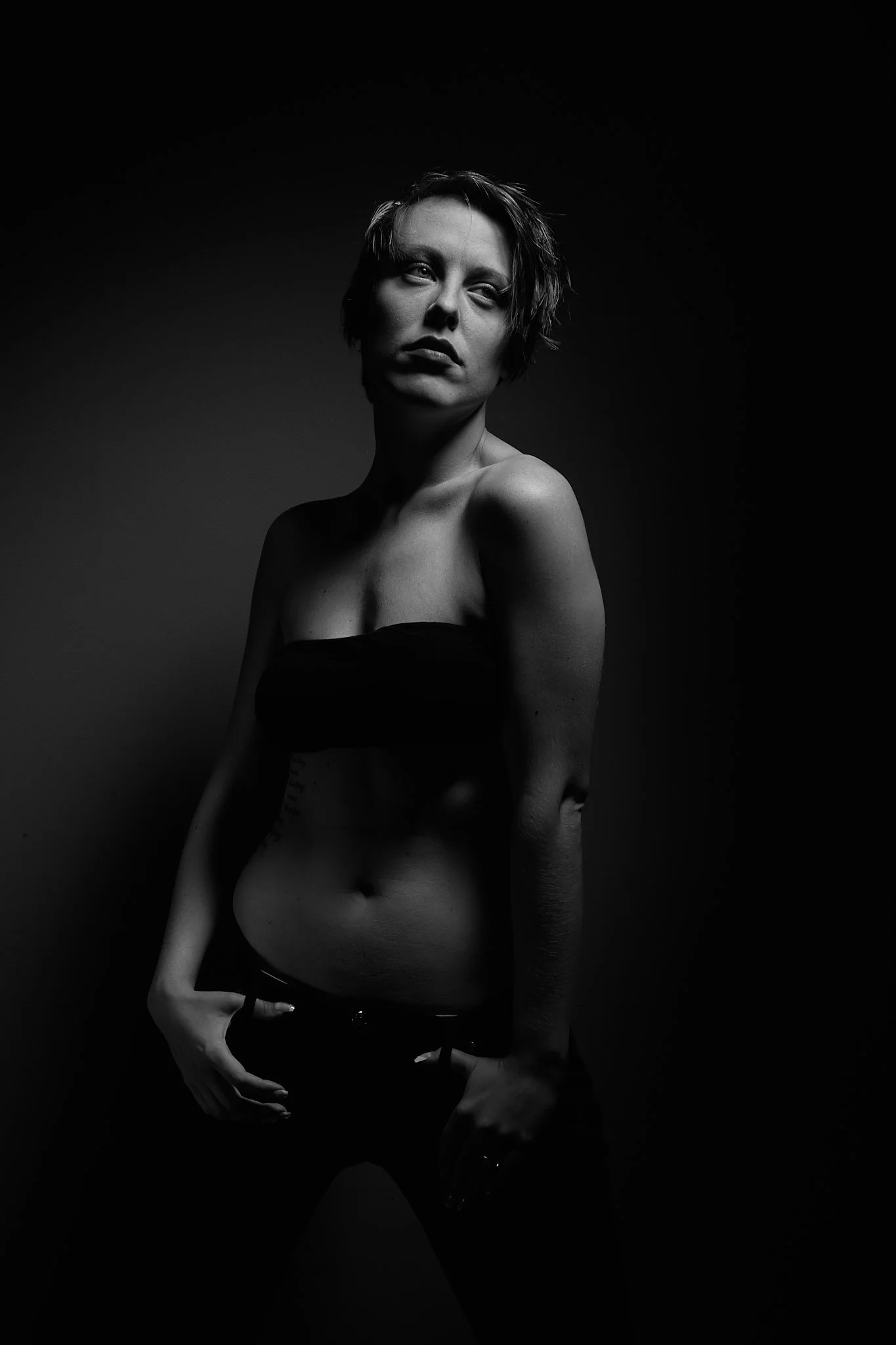 Black and white portrait of a woman with short hair, wearing a strapless top, standing against a plain dark background.