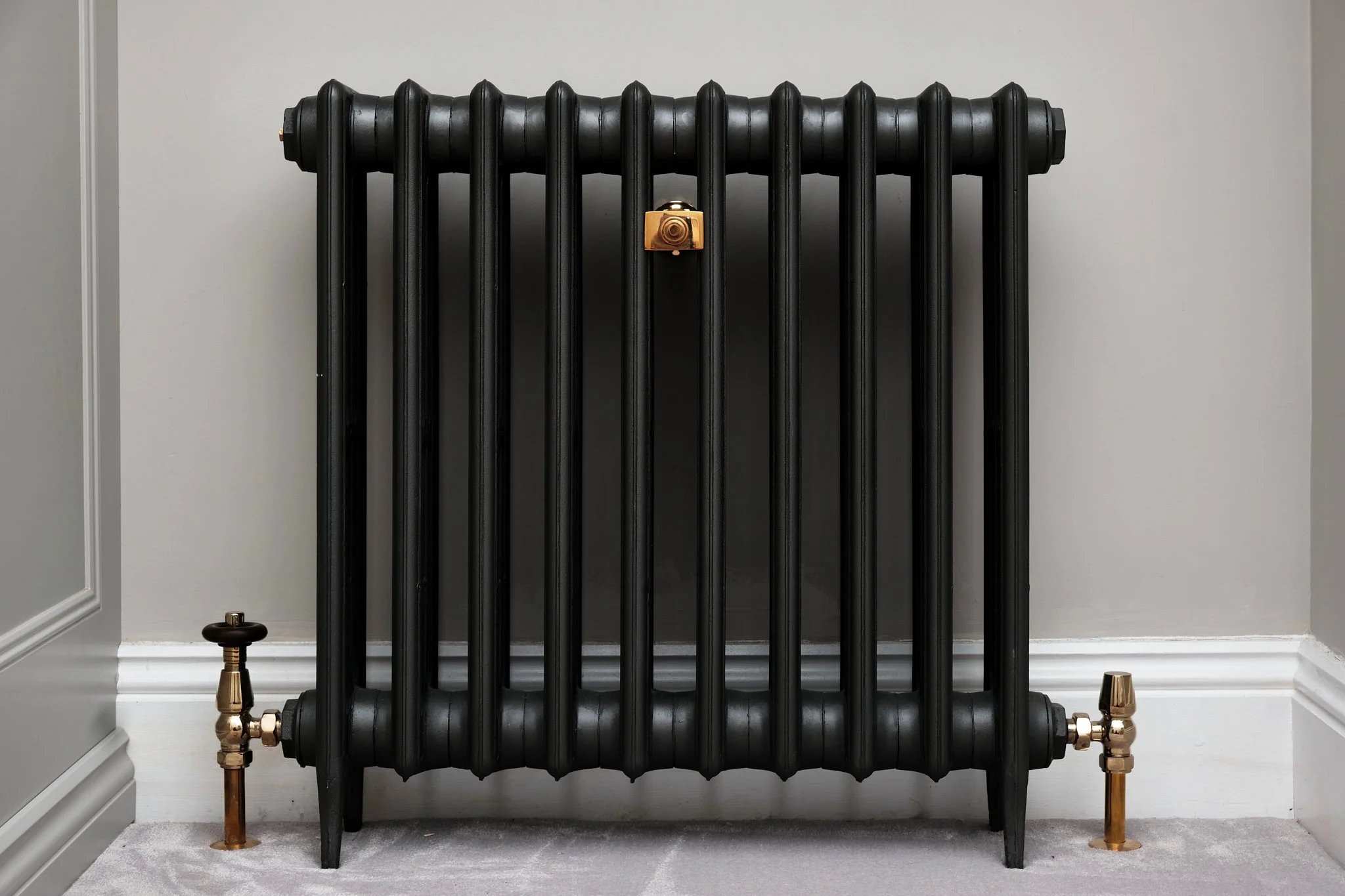 Black vintage radiator with brass fittings, positioned against a light gray wall on a white carpeted floor.