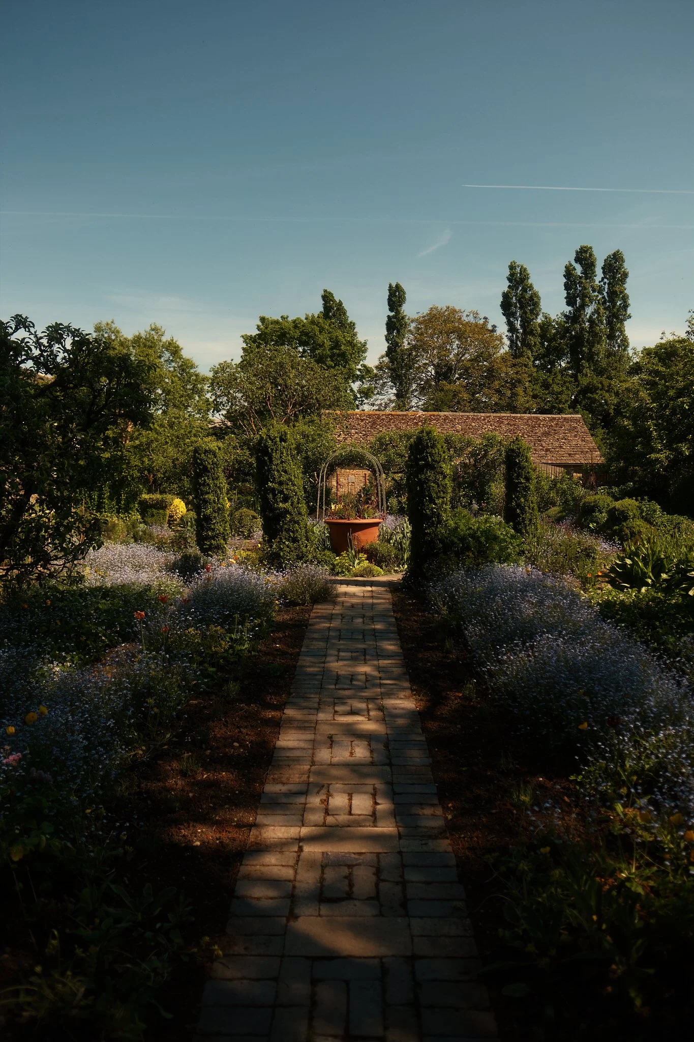 A garden pathway with trees and flowers leading to a small building and a large pot, under a clear sky. Hotel interior photography at THE PIG-in the Cotswolds