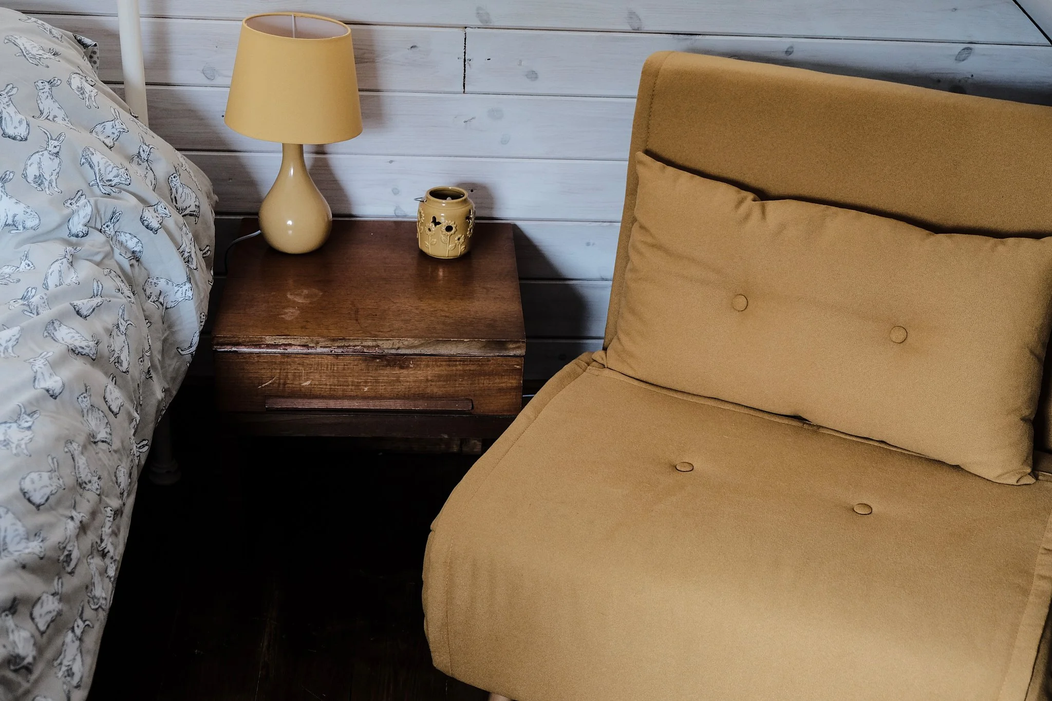 A bedroom with a bed on the left, a wooden nightstand, a beige lamp, a decorative jar, and a beige chair with a pillow. The wall behind is wooden paneling.