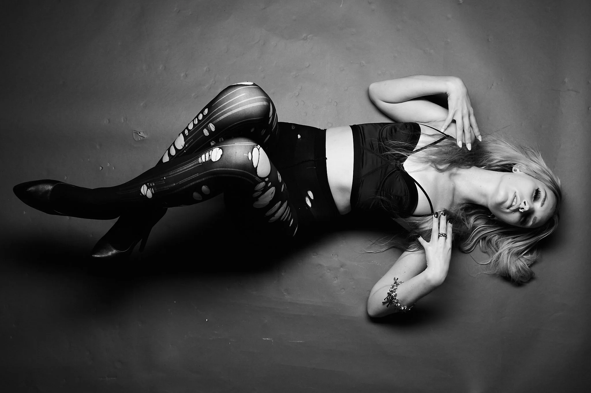 A woman with long hair lying on the floor, wearing a black top, patterned leggings, and high heels, in a black and white photo.