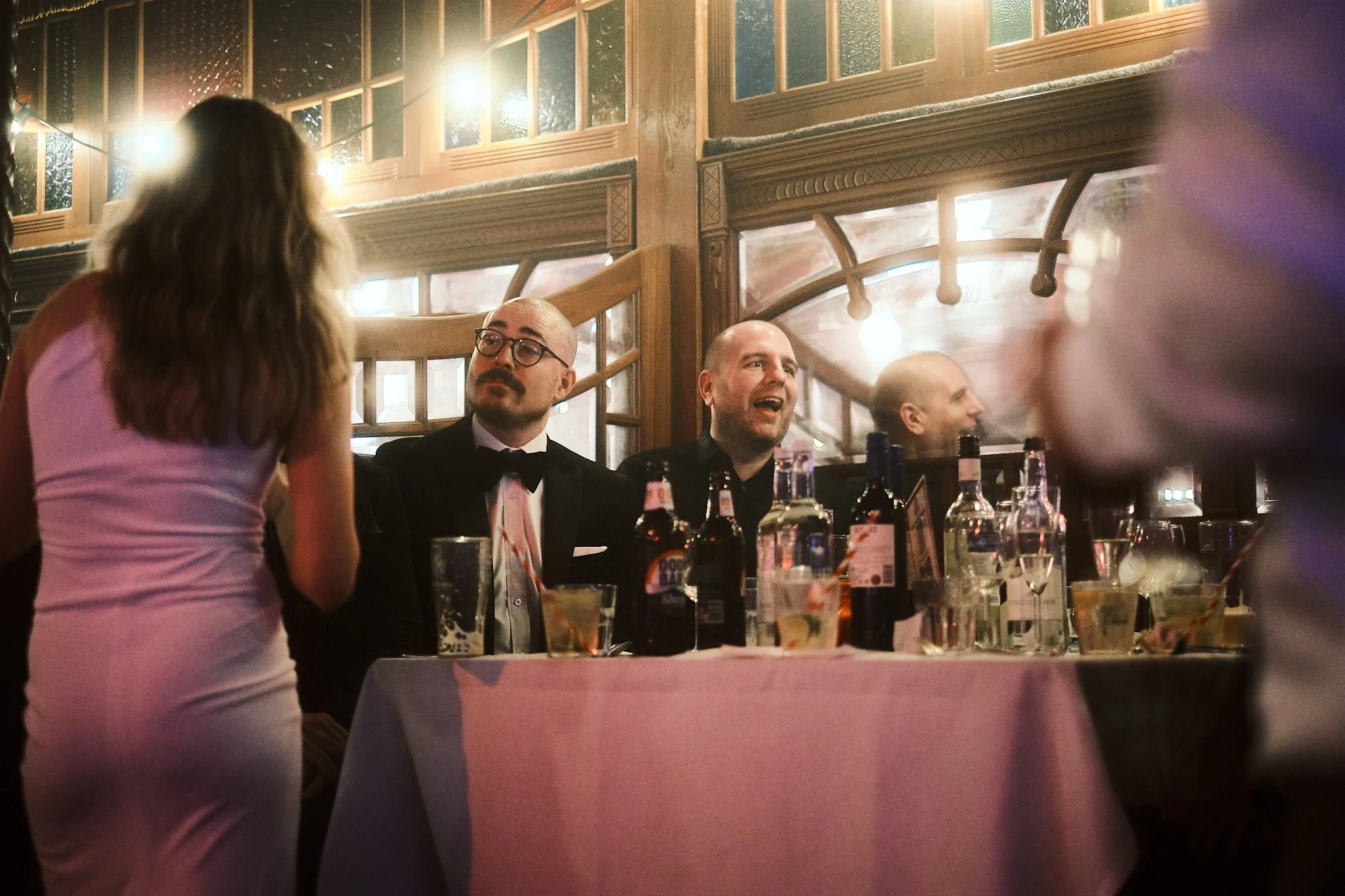 People sitting at a table in a restaurant or event space, engaging in conversation. The table has drinks including beer bottles, glasses of water, and some mixed drinks. The ambiance has warm lighting and wooden decor.