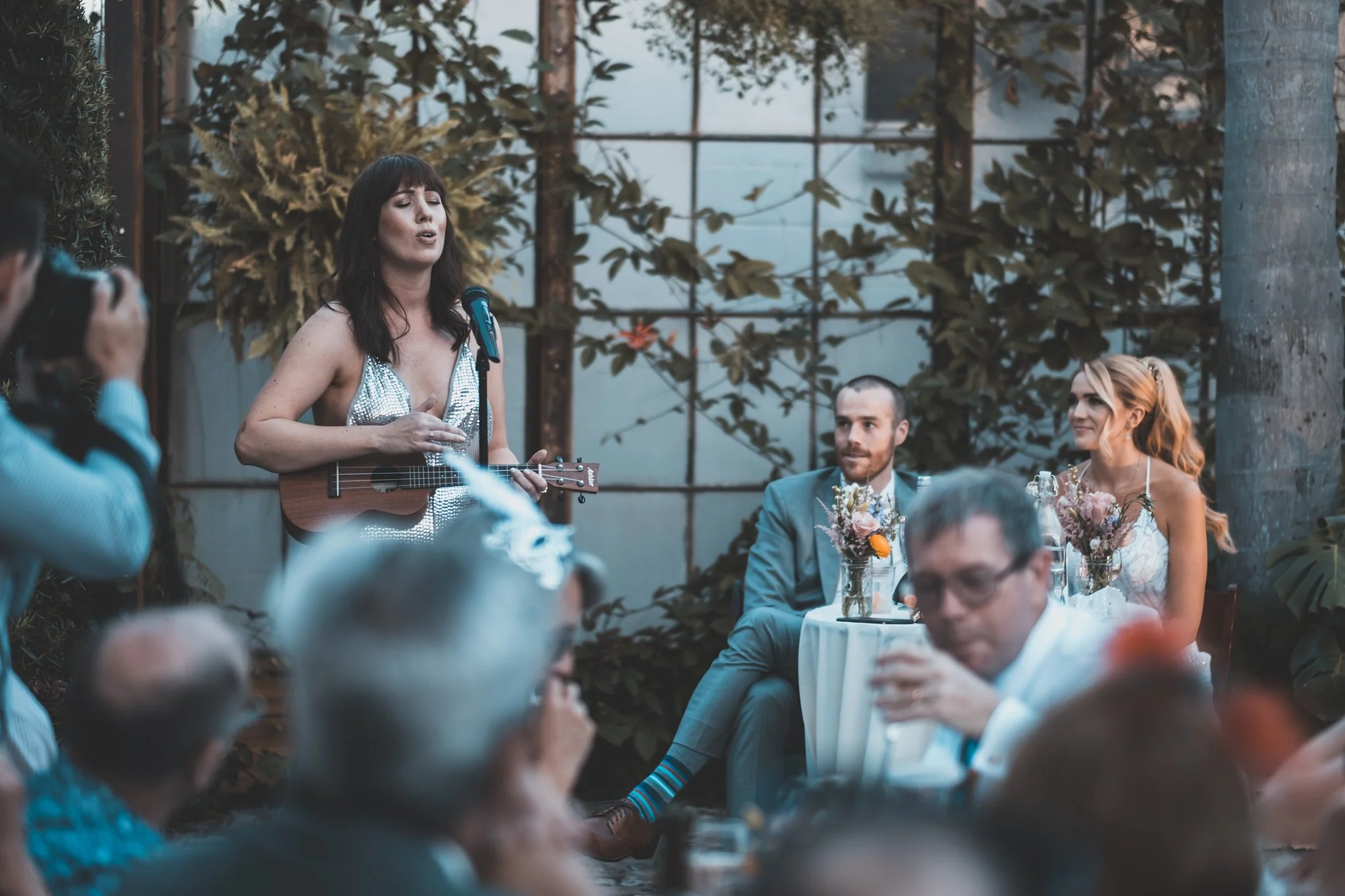 A woman playing a small guitar and singing at a wedding reception, with guests seated at tables around her. The scene is set in a greenhouse with plants and large windows.