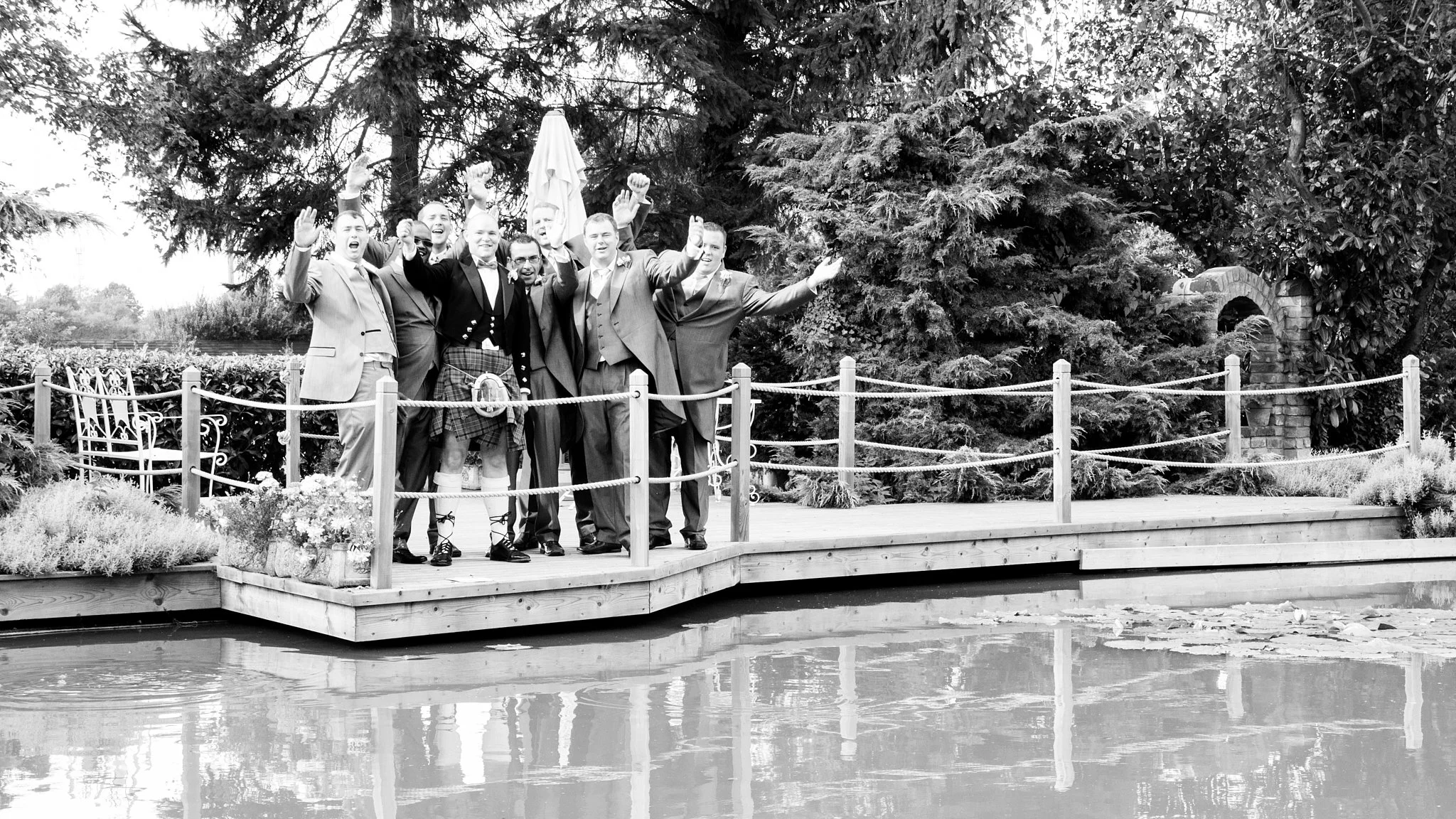 Men in formal attire celebrating on a wooden platform surrounded by trees and water, some with fists raised, in black and white.
