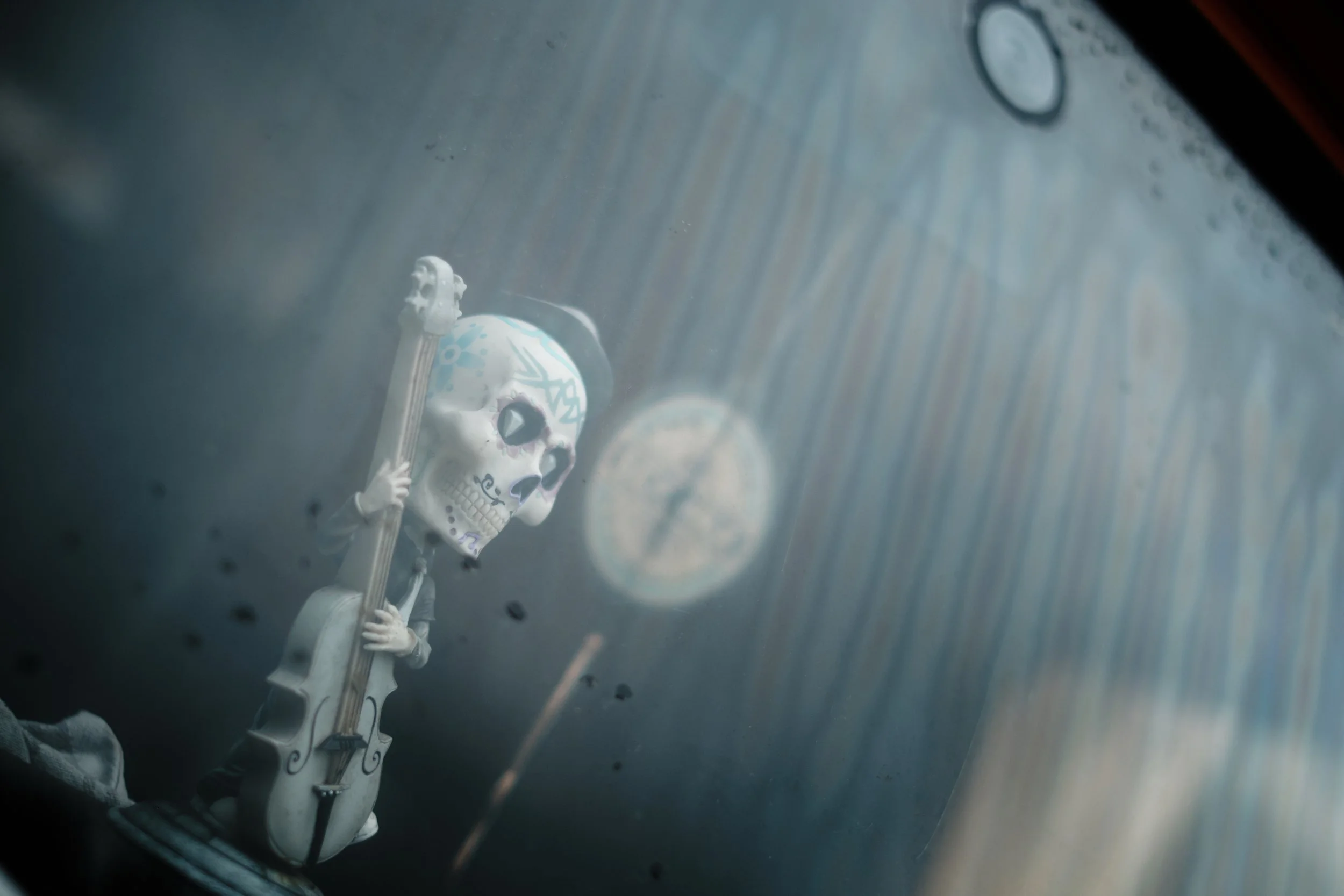 A small skeleton figurine with decorated skull is playing a guitar. The figurine has Day of the Dead style face paint and is inside a glass container with water drops, next to a fish bowl with a clock inside.