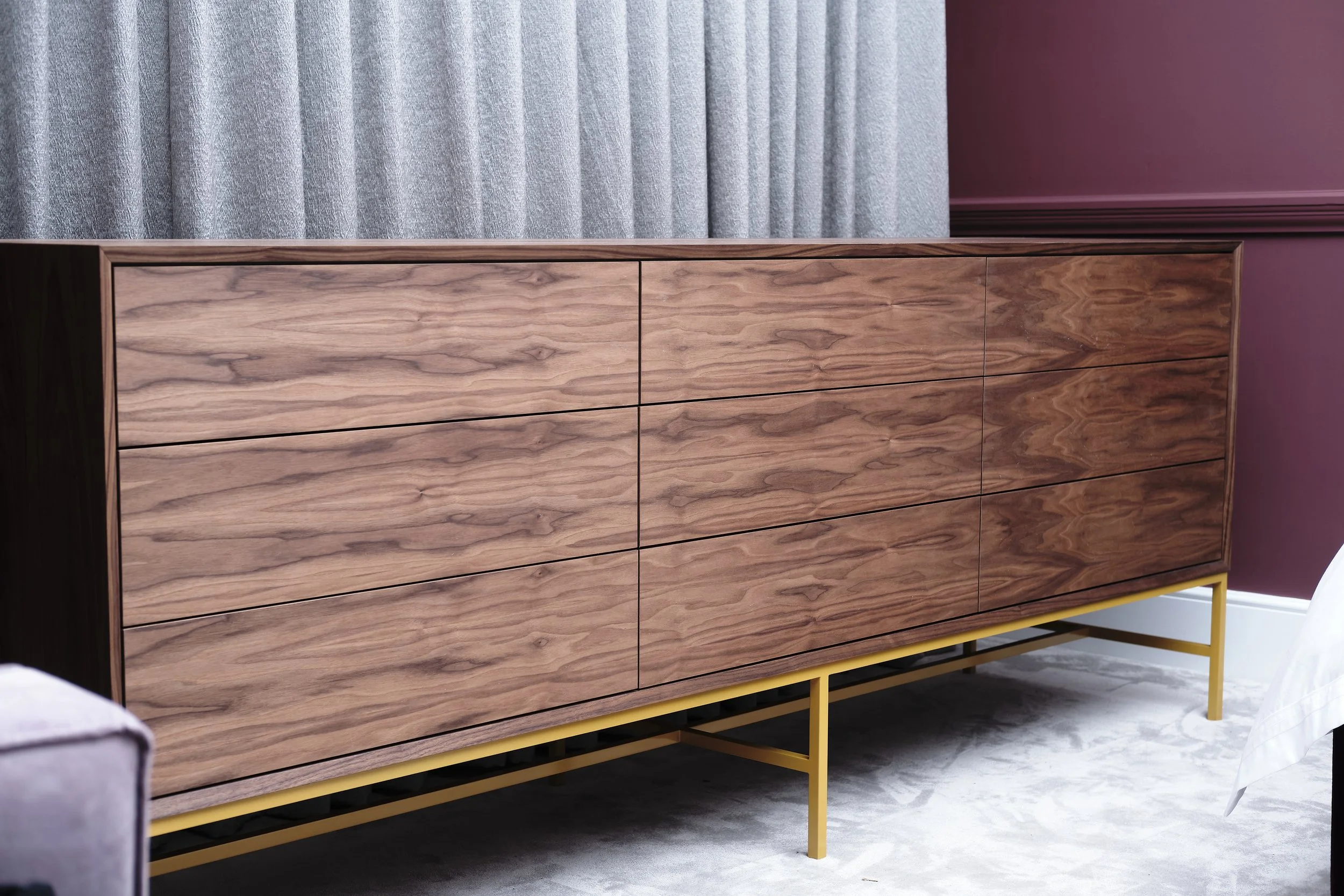 A wooden dresser with six drawers and a yellow metal base, positioned against a wall with purple paint and grey curtains.