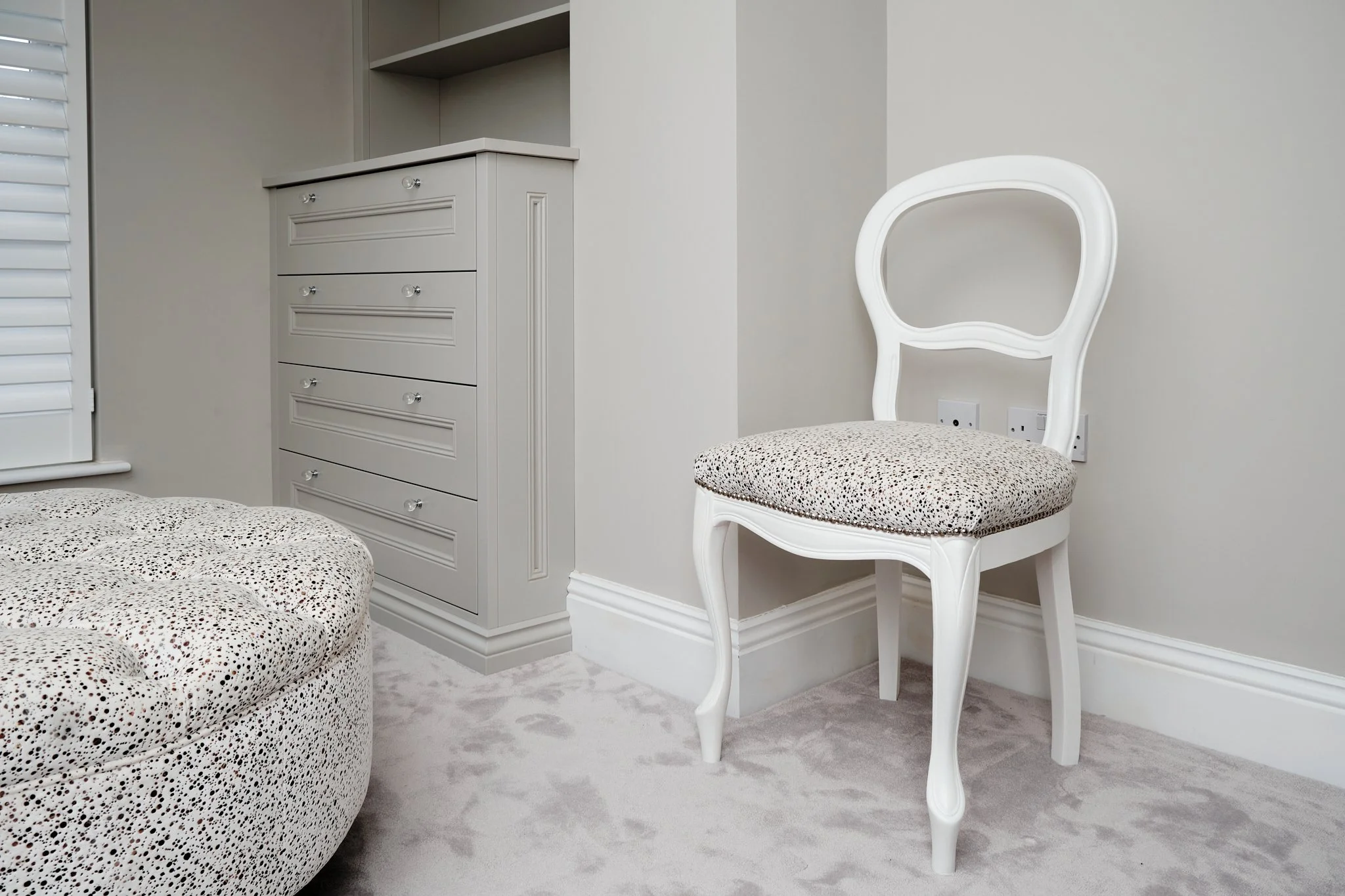 A white chair with an oval back and cushioned seat near a light gray wall, next to a dresser and a corner with a window.