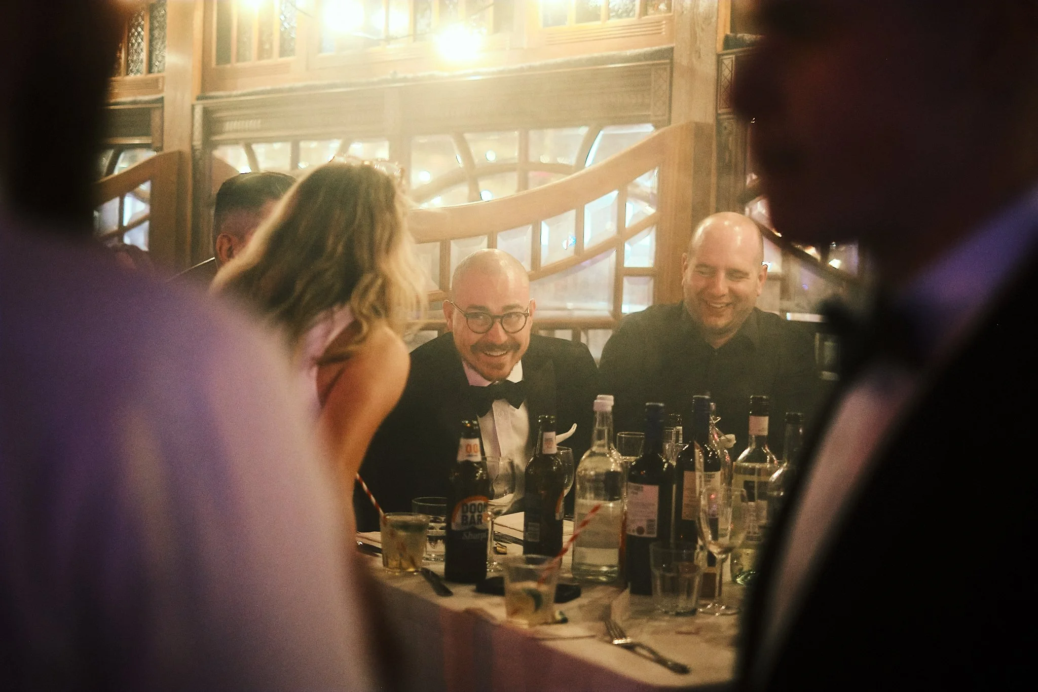 A group of people at a formal dinner, smiling and talking, with a man in a tuxedo and bowtie in the center, surrounded by drinks and bottles on the table.