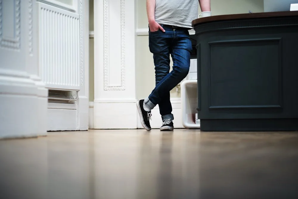 Person in jeans and sneakers leaning against a kitchen counter with a hand in pocket.