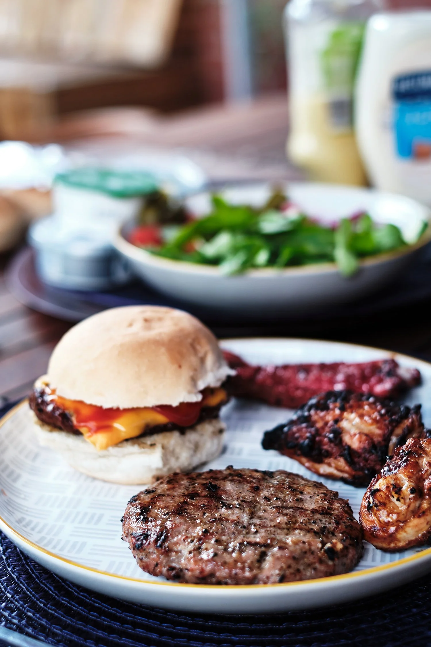 Close-up of a plate with a cheeseburger, grilled burger patty, bacon, and sausage, with a side of salad in the background.