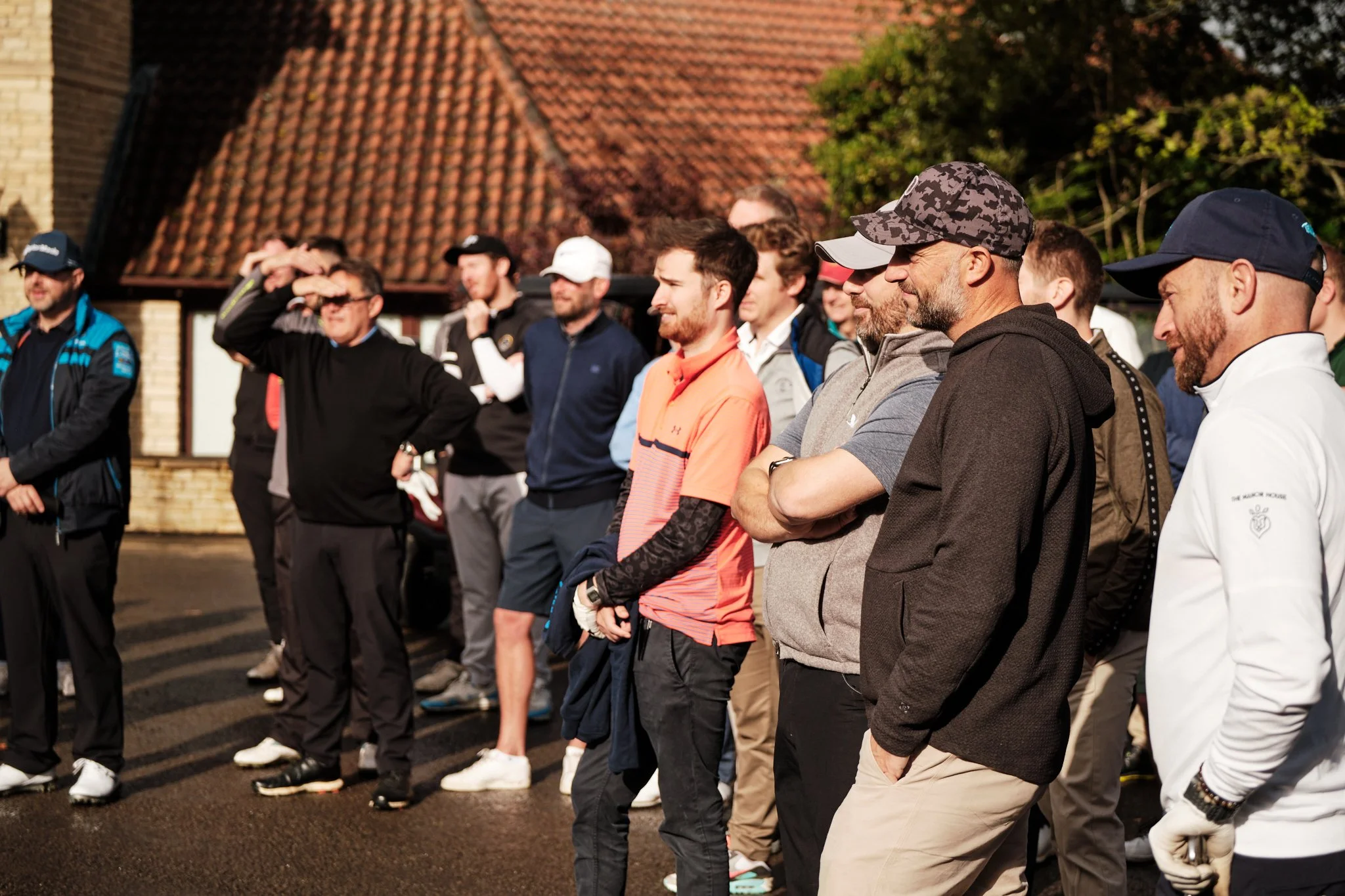 A group of people standing outdoors in front of a building with a tiled roof, dressed in casual sporty clothing, some wearing hats and sunglasses, possibly at a sporting or social event.