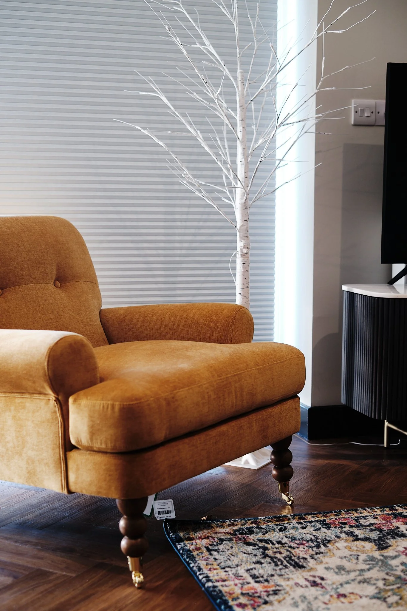 Living room with mustard-colored armchair, white decorative tree, black cabinet, on dark wooden flooring, with partially visible patterned rug.